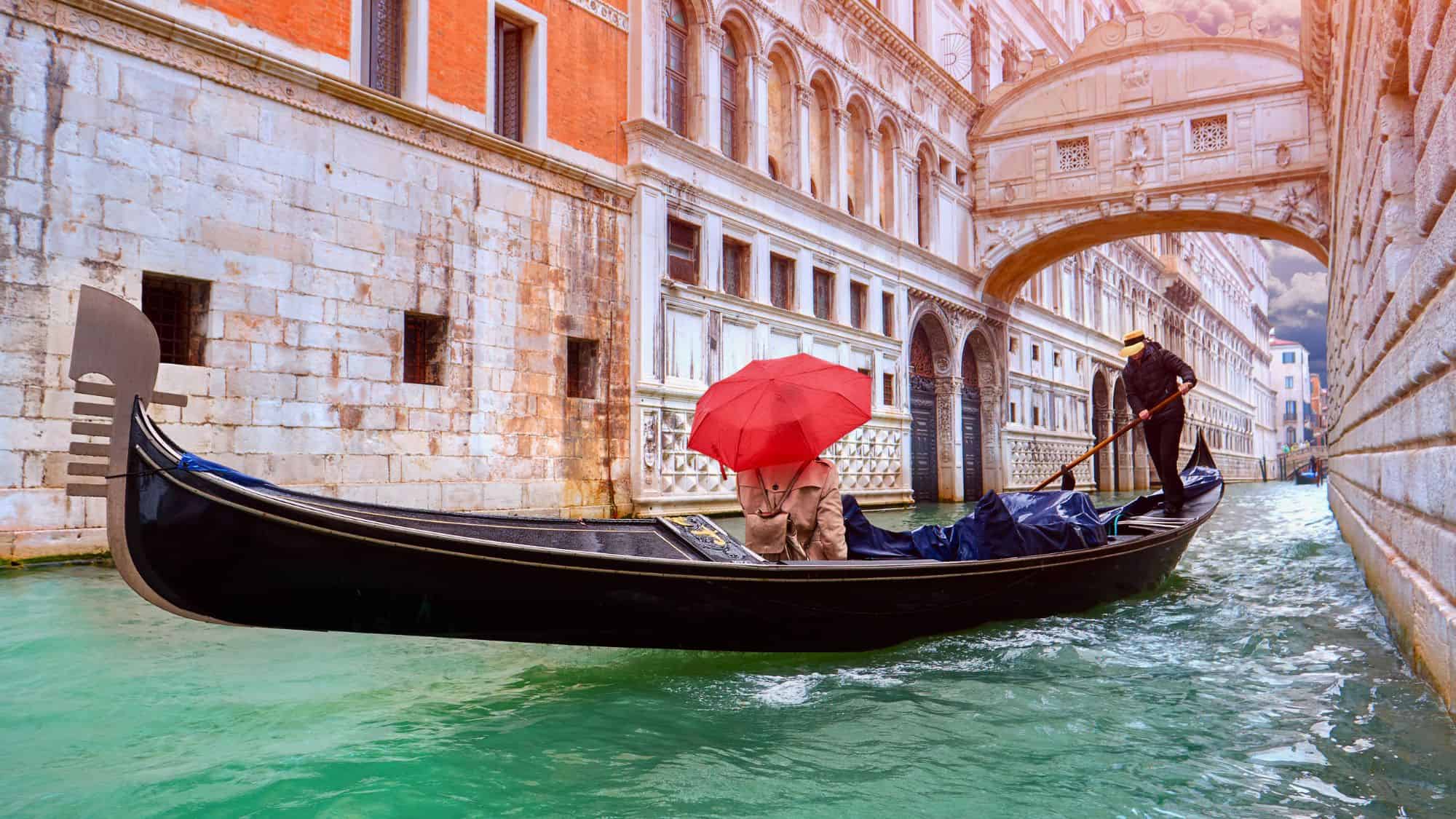 A gondolier navigates a narrow Venetian canal under the Bridge of Sighs while a passenger in a trench coat shelters beneath a striking red umbrella, adding a pop of color to the historic scene.