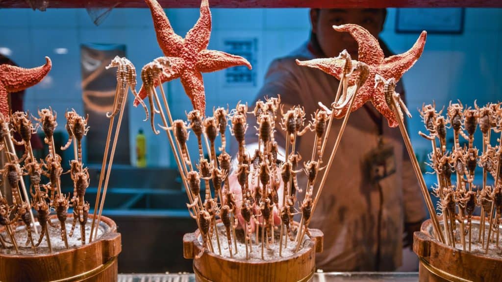 Close-up of exotic street food skewers at Wangfujing Snack Street in Beijing, featuring bright orange starfish and rows of scorpions on sticks, with a vendor partially visible behind the display.