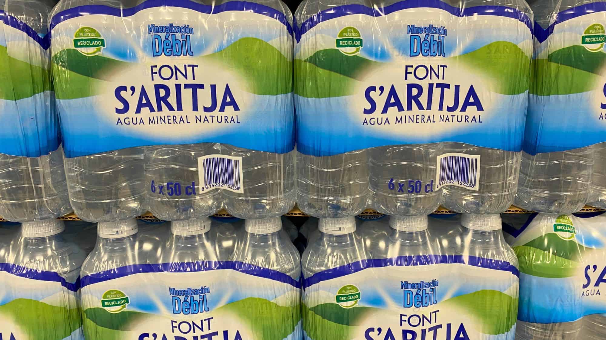 Shrink-wrapped packs of Font S’Aritja mineral water bottles stacked in a store, labeled “Agua Mineral Natural” with “Mineralización Débil” and “Con Plástico Reciclado” on the label.