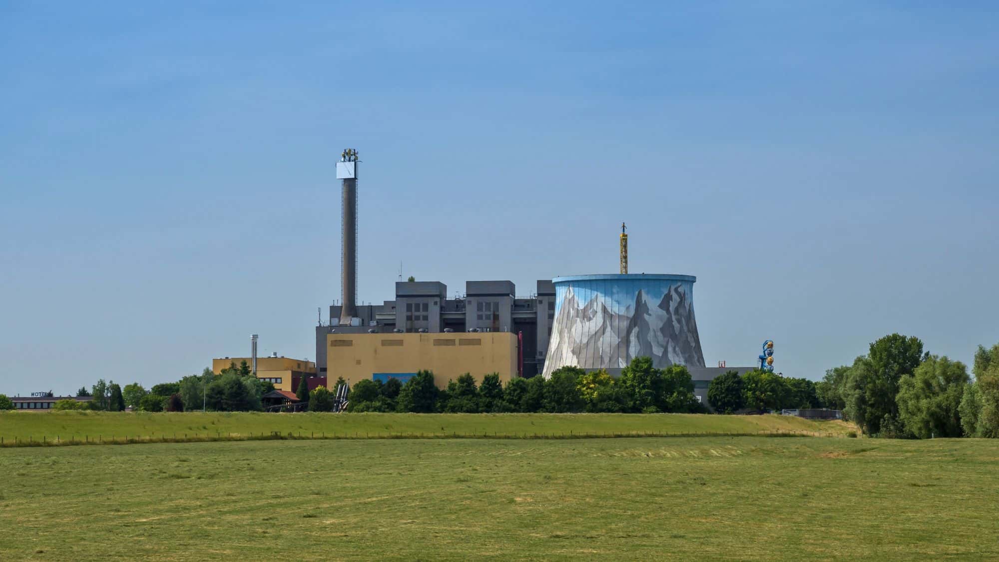 A wide view of the Wunderland Kalkar amusement park in Germany, showing an industrial cooling tower painted with mountain imagery and surrounded by modern buildings and green fields.