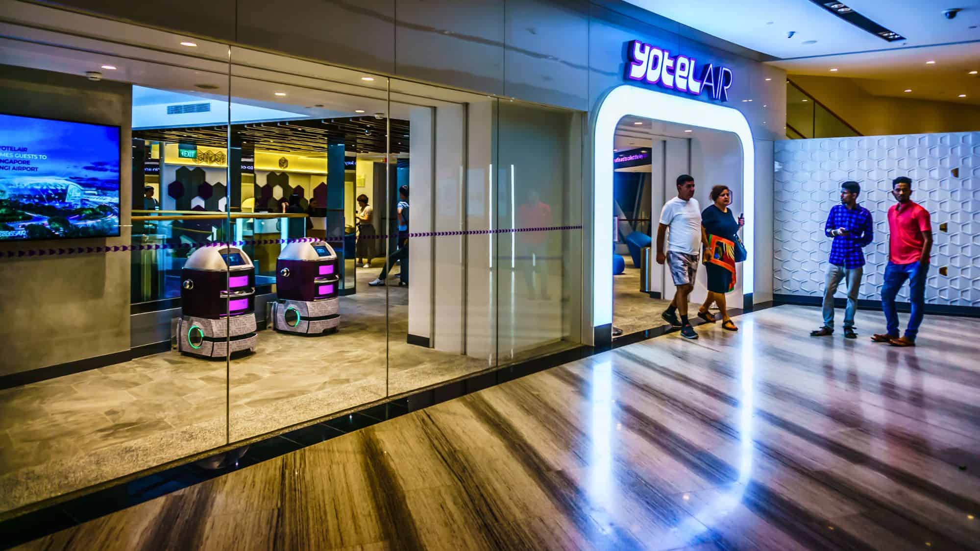 Brightly lit entrance to YOTELAIR at Changi Airport, featuring a sleek modern design with geometric wall patterns and two purple delivery robots near the glass doors as travelers pass by.