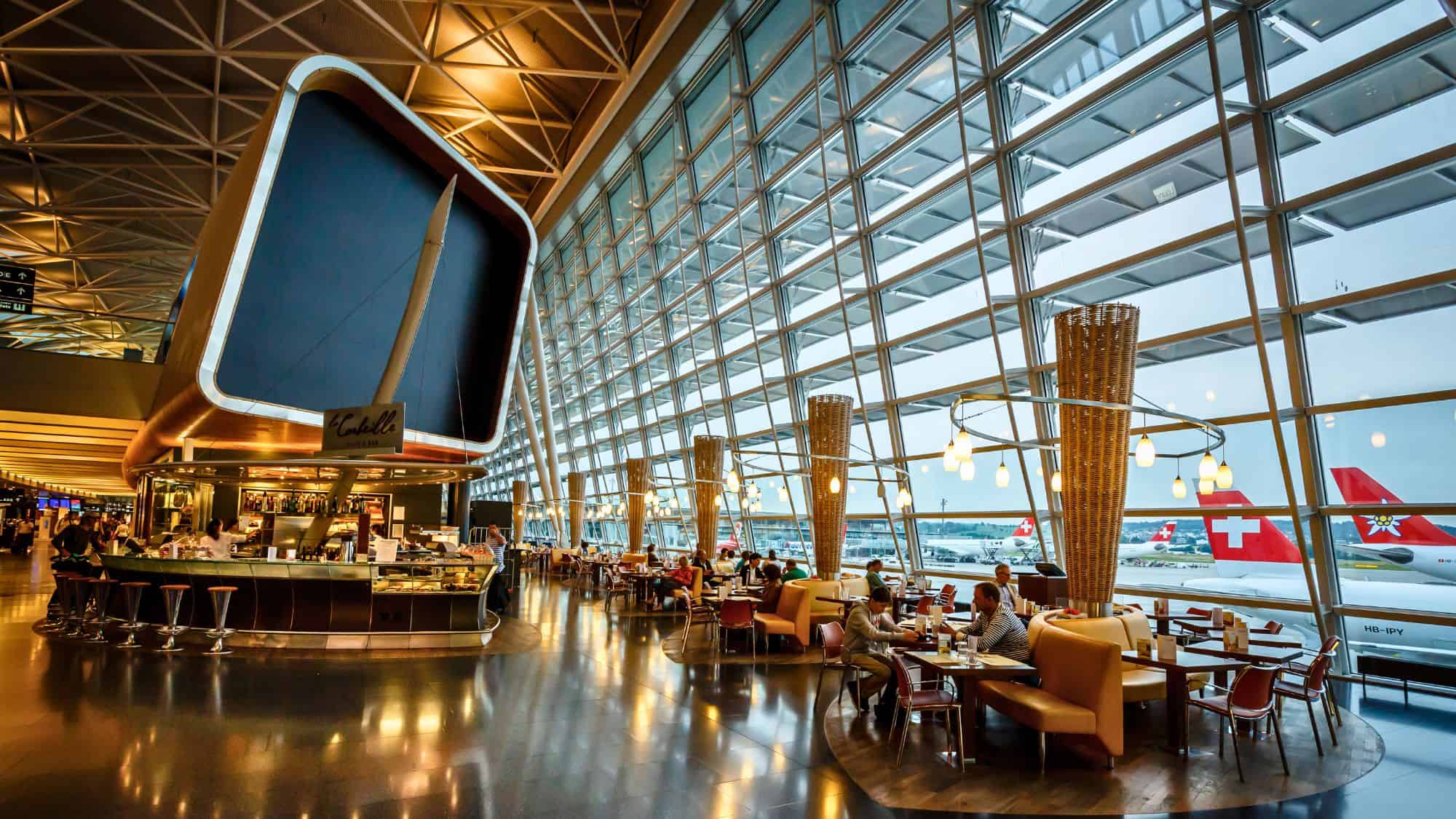 Travelers sit at cozy cafe tables inside Zurich Airport next to a floor-to-ceiling window wall, with Swiss International planes and the airfield visible in the background under a stylish, modern roof structure.