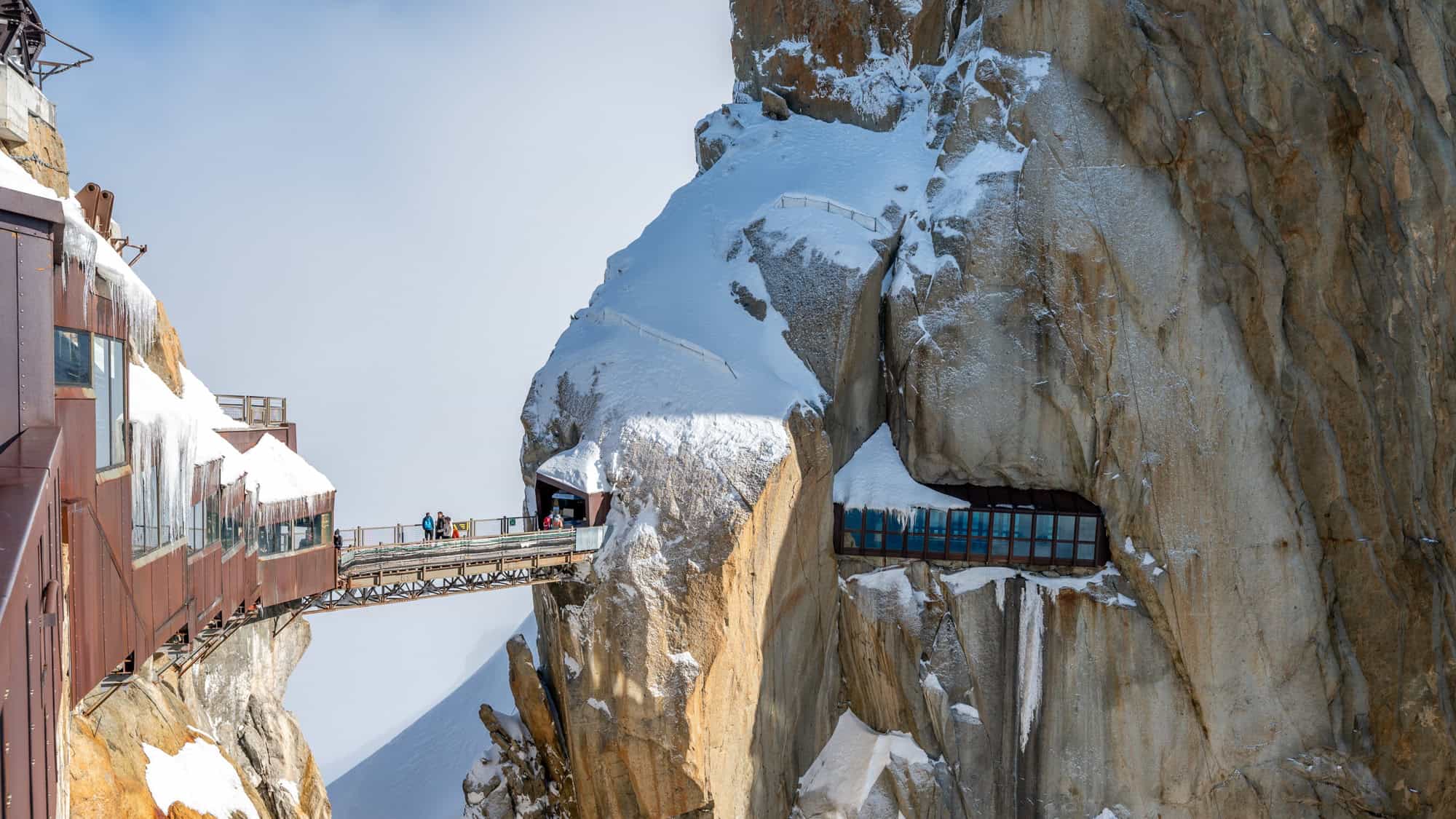 A narrow pedestrian bridge connects two snowy peaks at Aiguille du Midi in the French Alps, with modern red-and-glass buildings clinging to the cliffside amid snow and icicles.