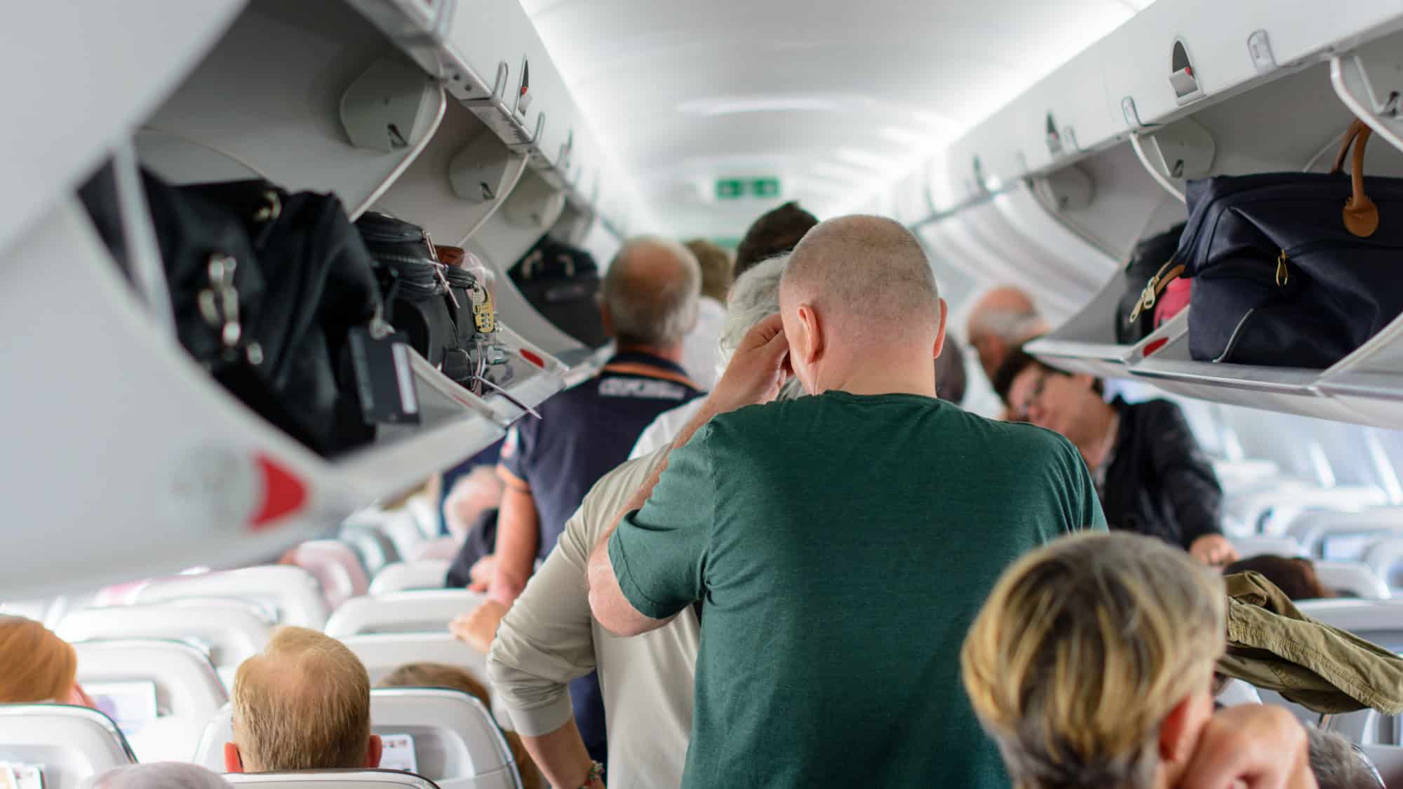 Passengers stand packed in the narrow aisle of a commercial airplane during boarding, reaching for overhead bins and waiting to be seated.