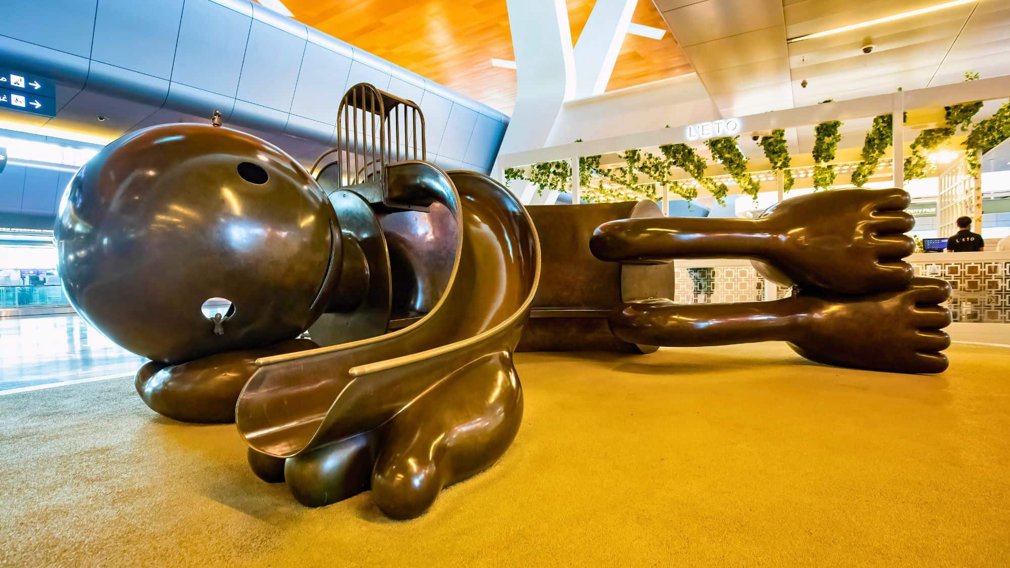 A whimsical bronze sculpture of a reclining cartoon character with exaggerated features, featuring a built-in slide as part of an interactive children’s play area in an airport.