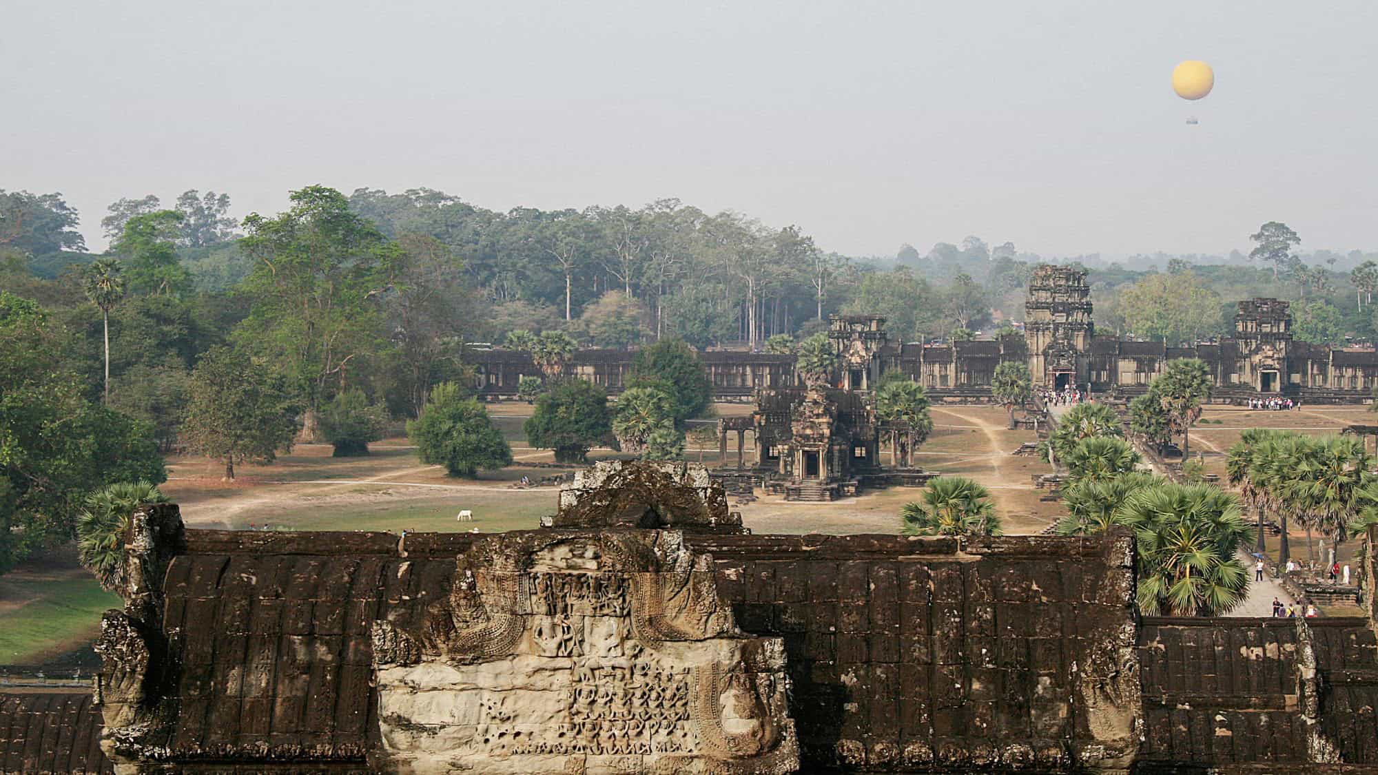 A yellow tethered hot air balloon floats above the ancient stone temples of Angkor Wat, Cambodia, surrounded by dense green forest and scattered palm trees.