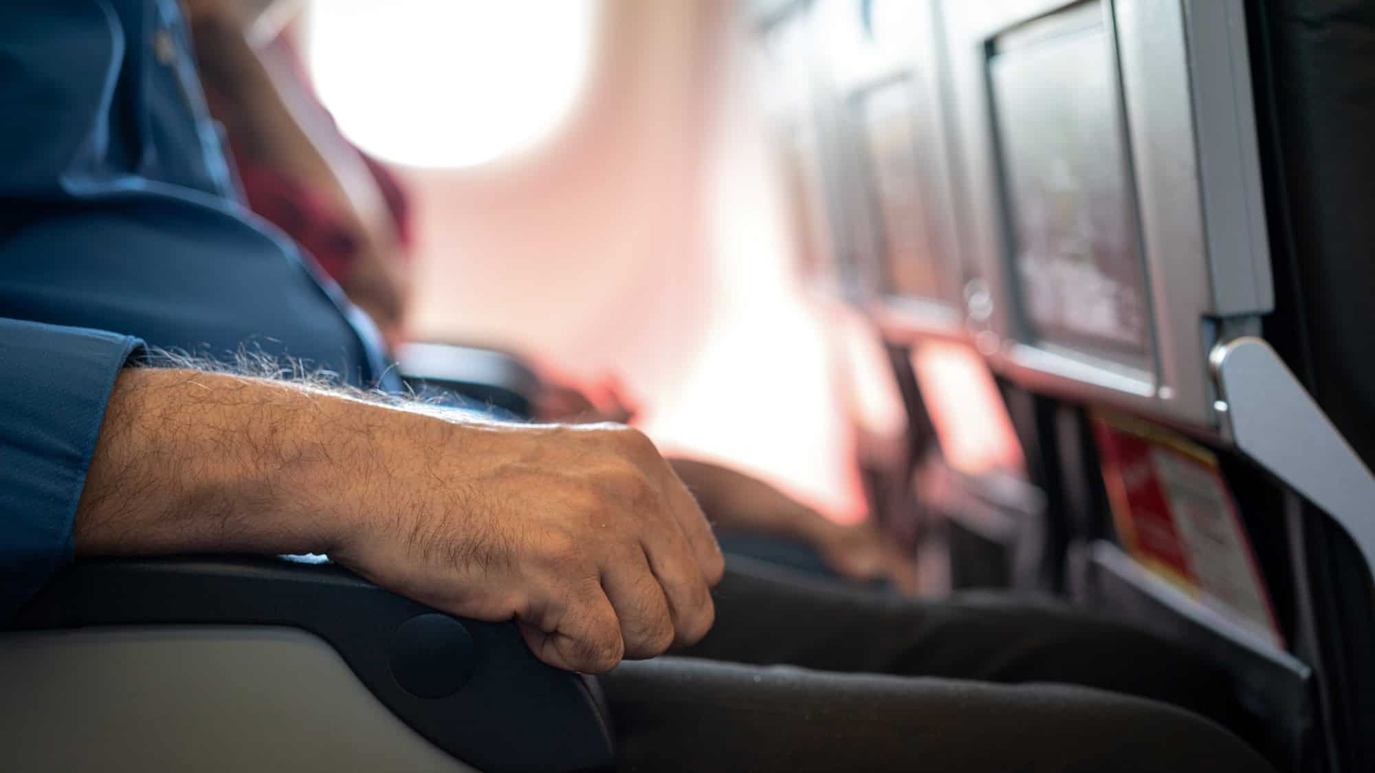 Close-up of a man’s hand gripping the armrest of an airplane seat, subtly conveying the territorial discomfort common in shared airplane seating.