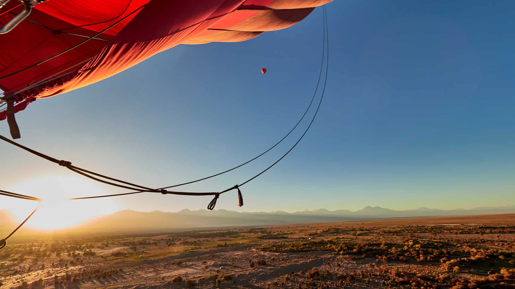 View from a hot air balloon ride over the Atacama Desert at sunrise, with the balloon’s red fabric and rigging in the foreground and another balloon floating in the distance.