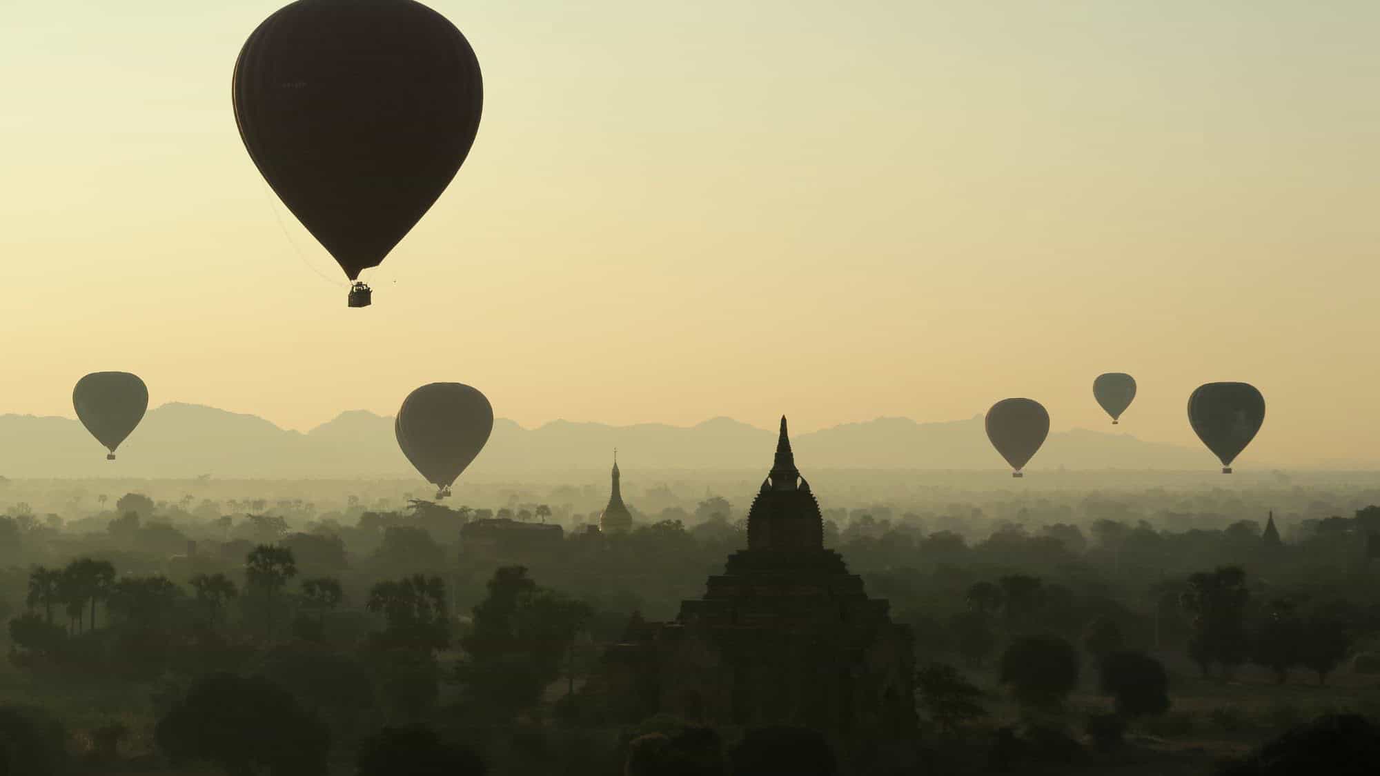 A hot air balloon floats above the ancient temples of Bagan, Myanmar, during a hazy sunrise. The reddish-brown pagodas peek through the mist across the flat plain, creating a dreamy, golden-lit scene.