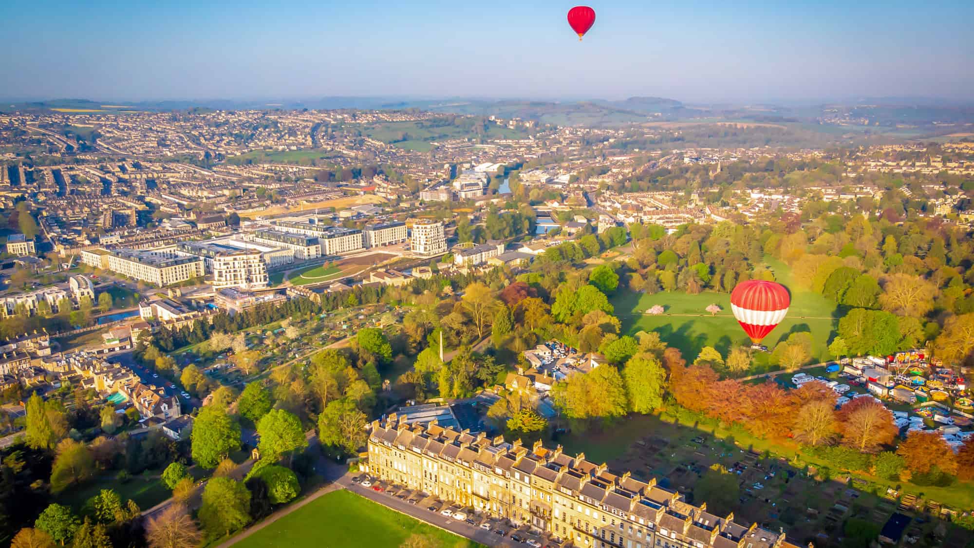 Hot air balloons soar above the historic cityscape and Georgian architecture of Bath, England, with manicured parks, stone buildings, and a clear morning sky.