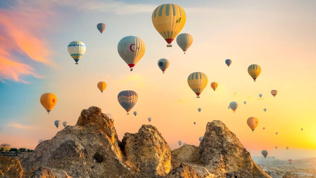 A sky filled with colorful hot air balloons rising above the rocky, otherworldly landscape of Cappadocia, Turkey, during a pastel sunrise that bathes the scene in soft orange and pink light.