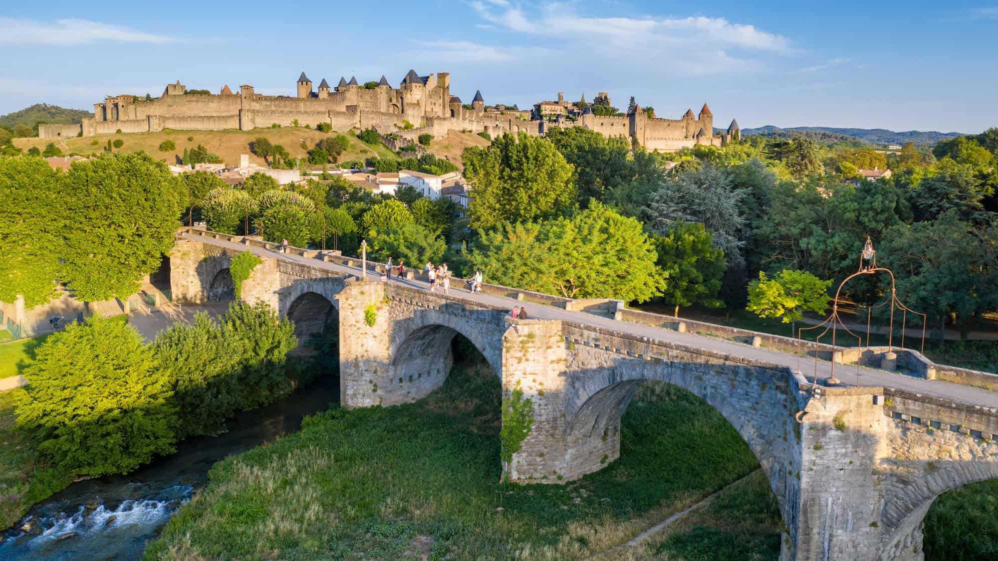 Impressive medieval walled city of Carcassonne in southern France with pointed turrets and fortified walls, viewed from a stone bridge crossing a tranquil river below.