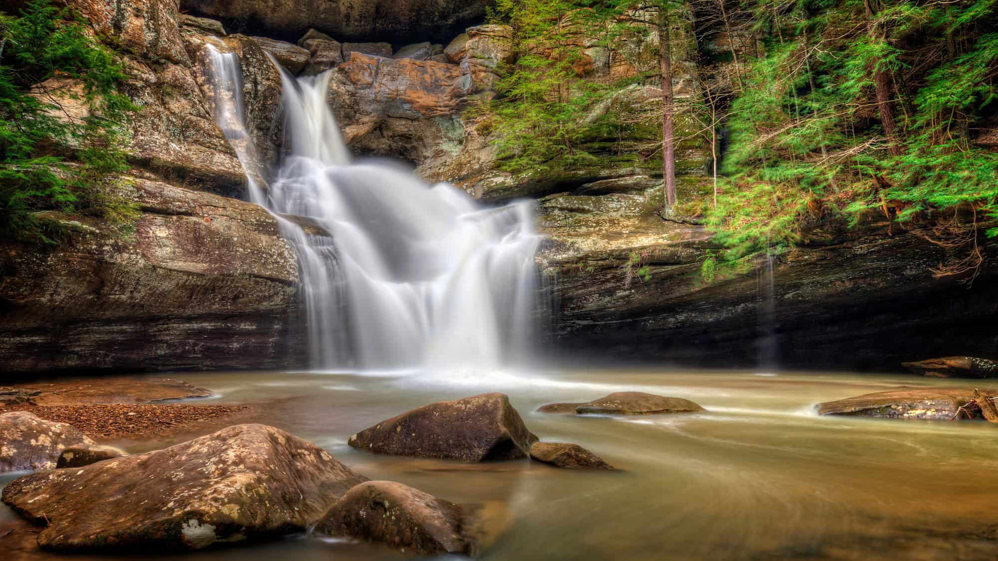A serene waterfall spills into a calm pool surrounded by mossy rocks and lush forest in Hocking Hills, Ohio, with smooth long-exposure water flow.