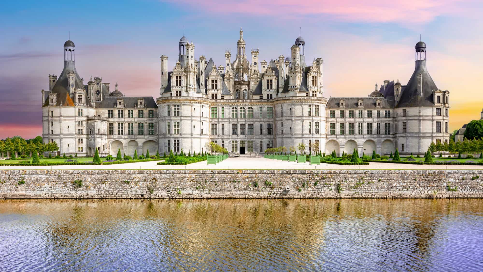 A grand Renaissance château in the Loire Valley with distinctive French towers and elaborate spires, reflected in a calm moat under a soft pink and purple sunset sky.