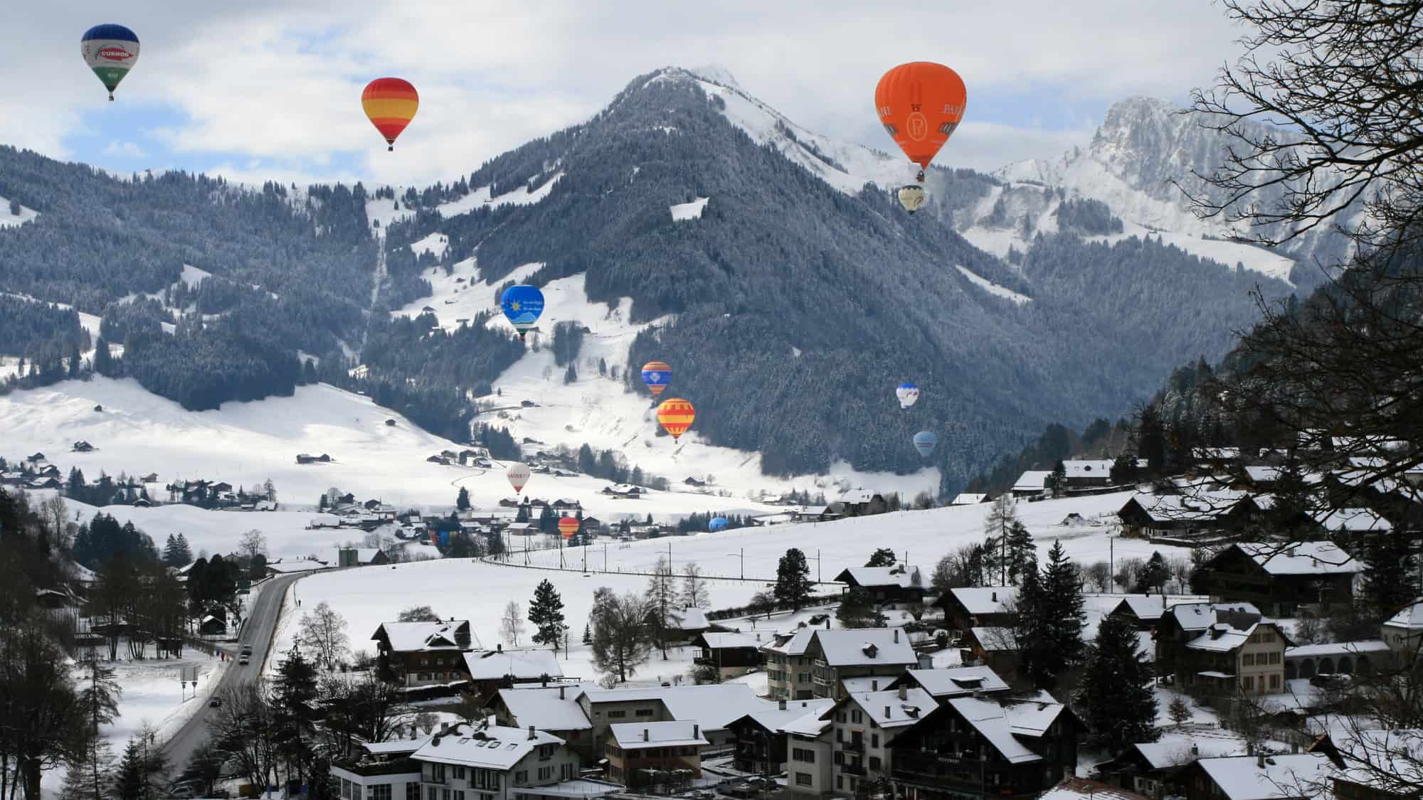 A colorful mix of hot air balloons rises above a snowy Swiss alpine village during winter, with frosted trees and steep mountain slopes in the background.