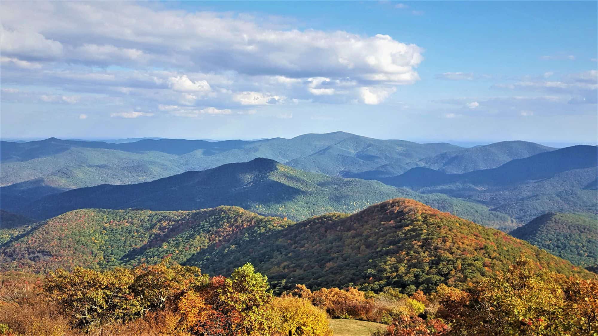 A panoramic view of rolling, forested mountains blanketed in early autumn colors under a bright blue sky with scattered clouds, showcasing the vast, layered ridgelines of northern Georgia.