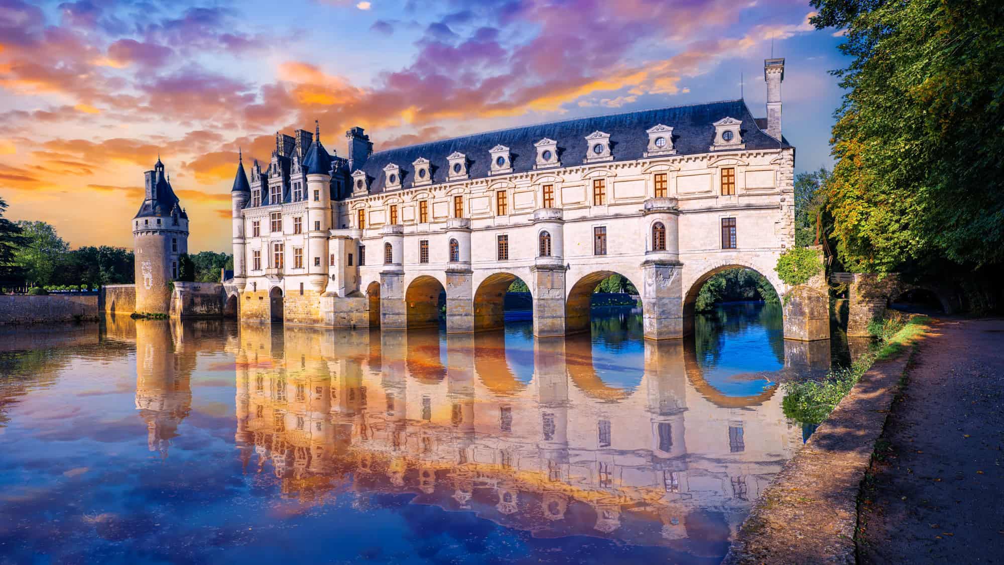 The fairytale Château de Chenonceau arches over the River Cher, its pale stone façade mirrored in the water below during a dramatic, colorful sunset.