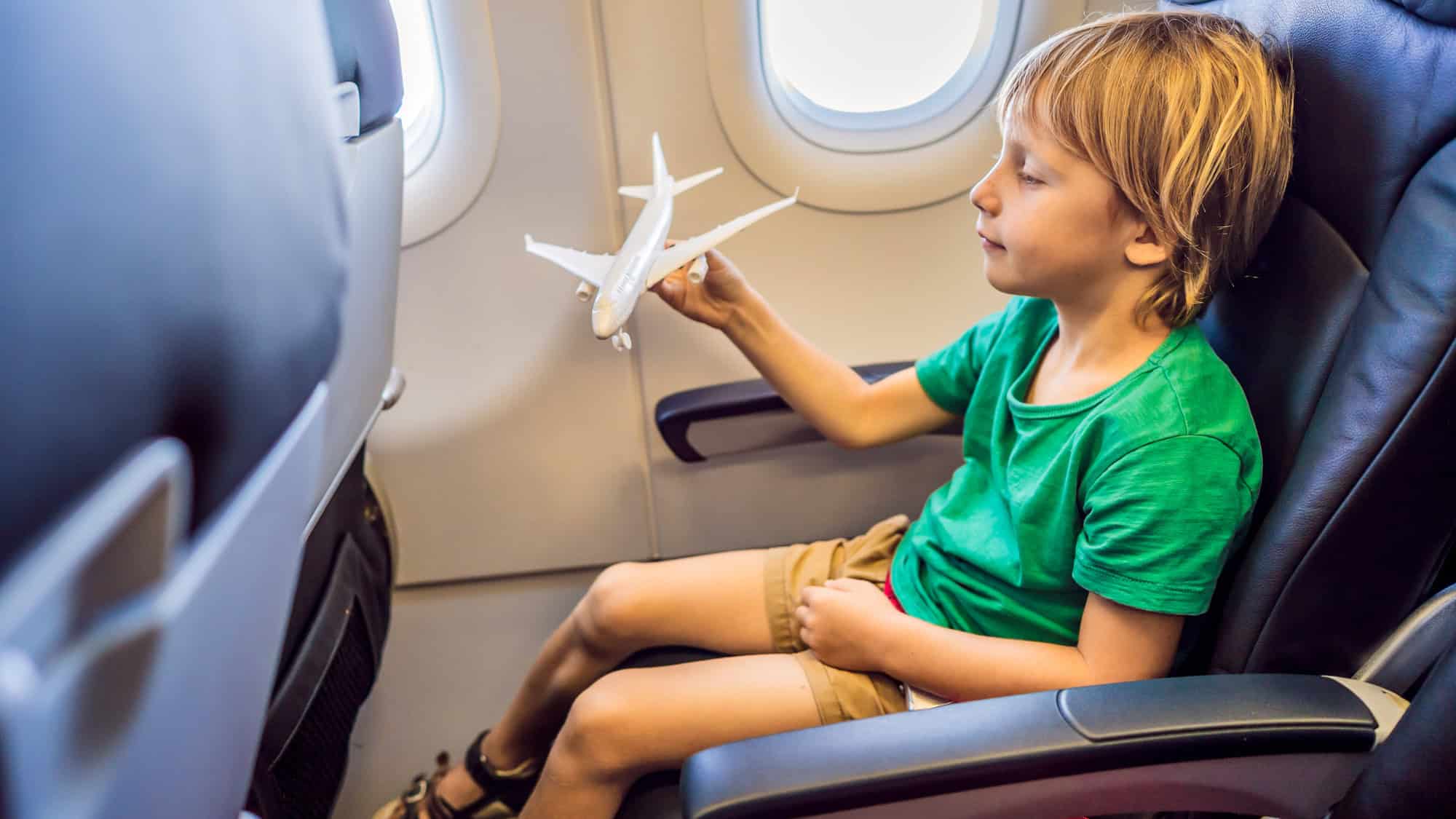 Young boy in a green shirt and tan shorts sits in an airplane seat holding a toy airplane, looking amused as he imagines it in flight.