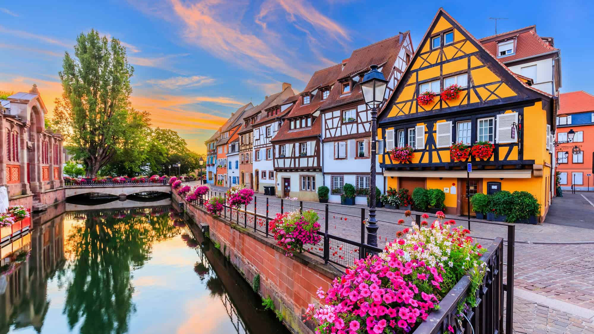 Colorful half-timbered houses line a peaceful canal in Colmar, France, with vibrant flower boxes overflowing onto a cobblestone path at sunset.