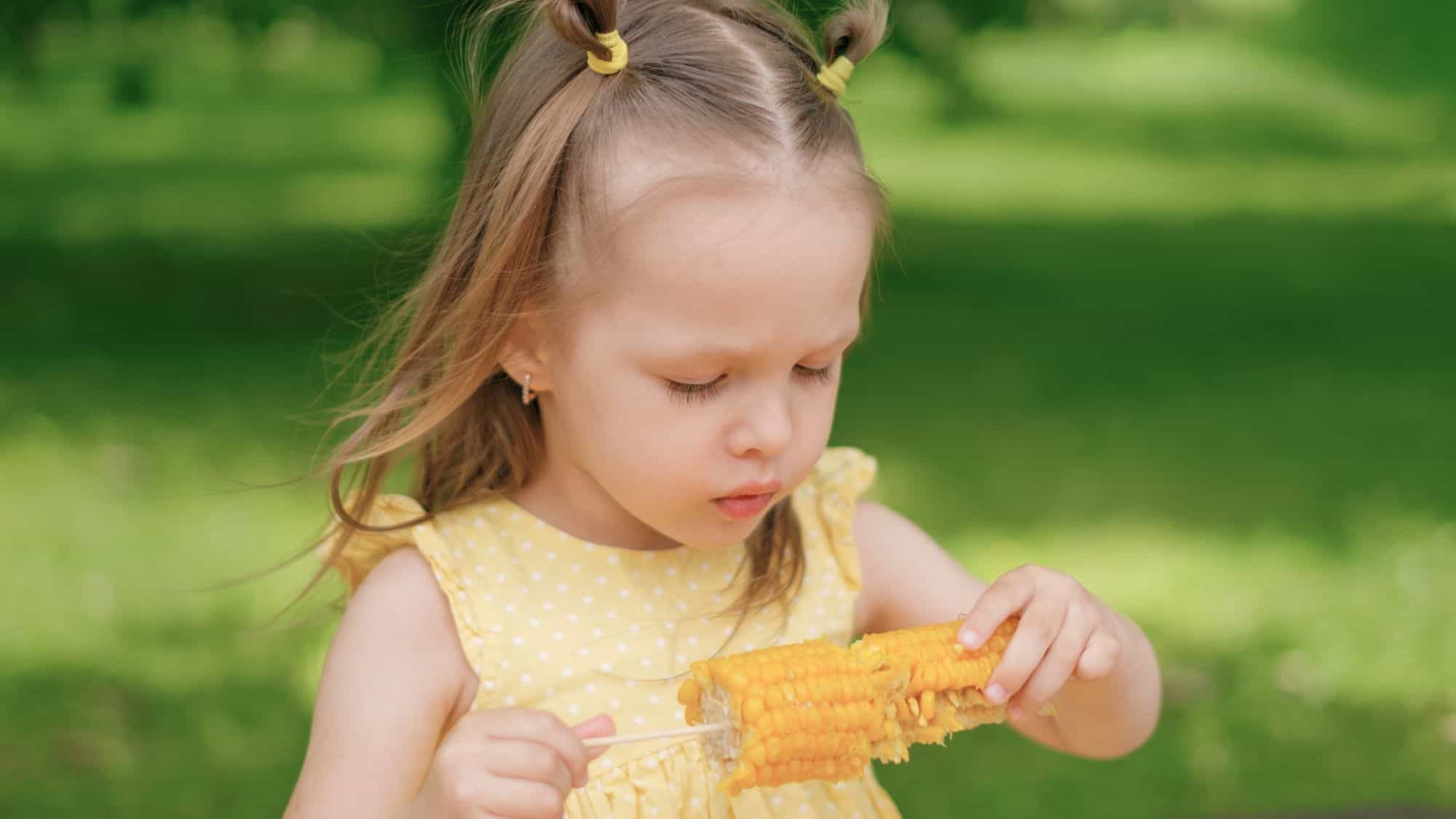 A young girl in a yellow polka-dot dress concentrates on eating corn on the cob outdoors, with a lush green background.