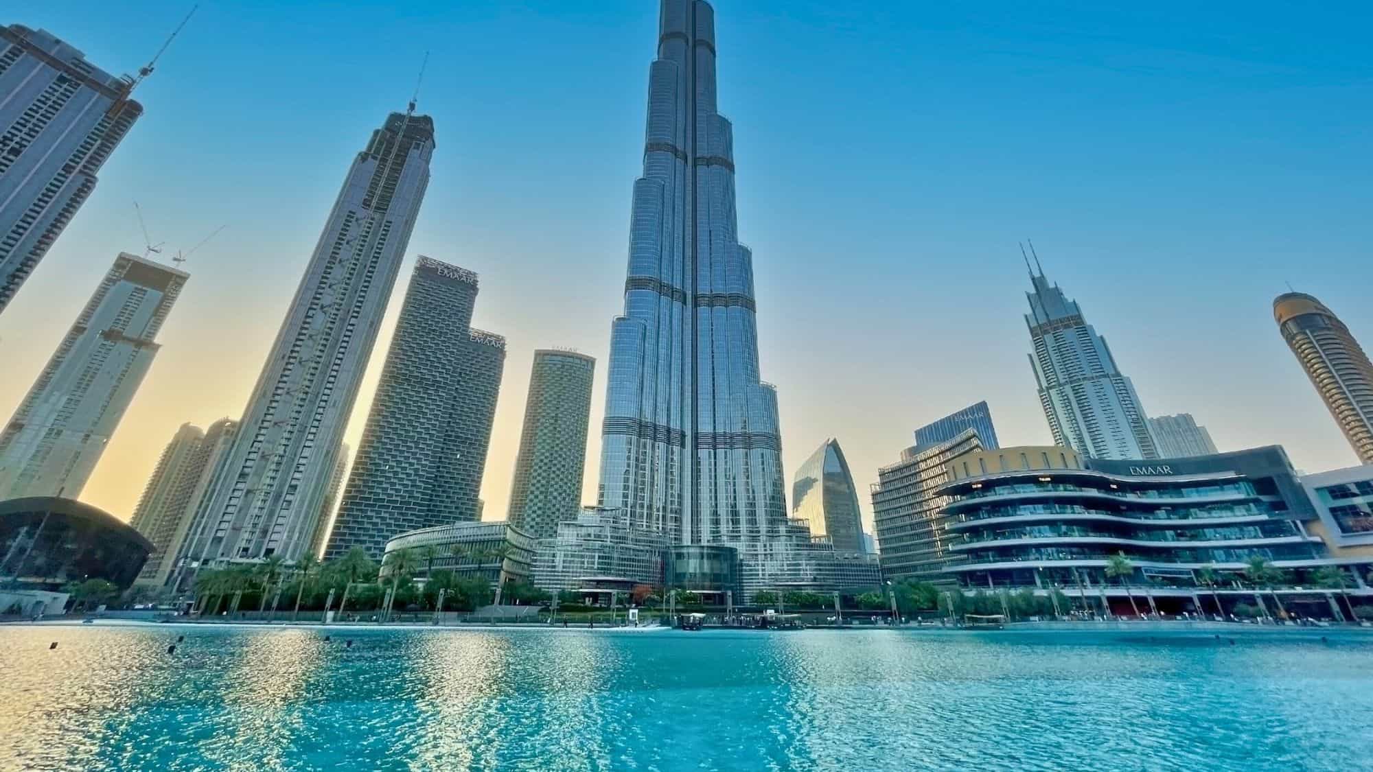 Wide-angle view of the Burj Khalifa in Dubai rising high above surrounding modern skyscrapers, with its glassy surface reflecting the soft golden glow of sunset. A large turquoise fountain pool spans the foreground, adding contrast to the sleek urban scene.