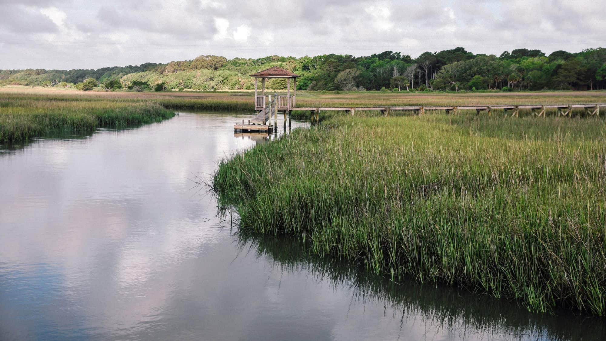 A peaceful marshy inlet on the Edisto River in South Carolina, featuring tall green grasses, a wooden dock with a gazebo, and a cloudy sky overhead.