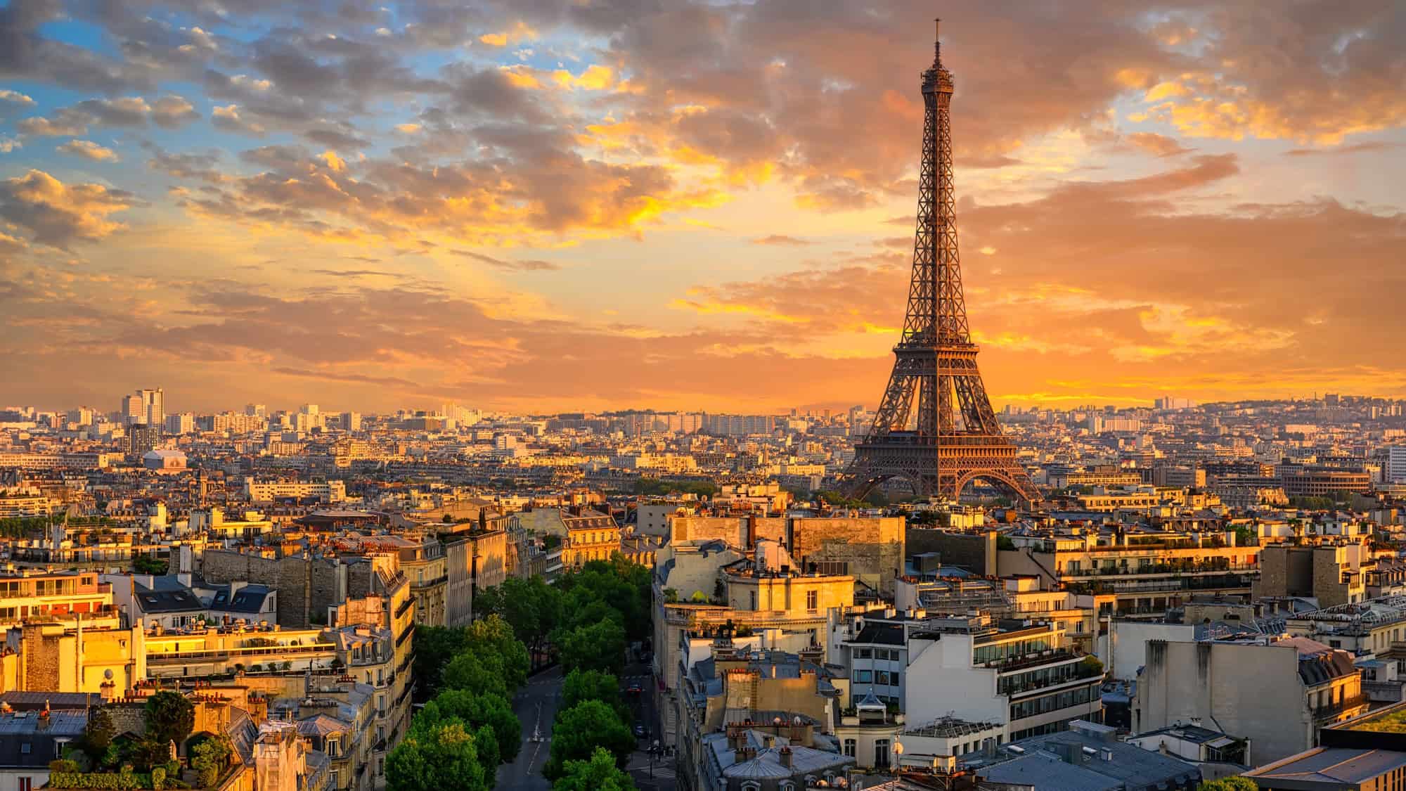 The Eiffel Tower stands tall over Paris as the sun sets, casting golden light over the rooftops and cityscape below, with scattered clouds adding drama to the glowing sky.