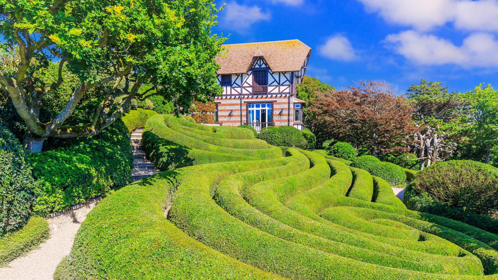 Charming timber-framed house at the top of Etretat’s cliffside gardens, surrounded by meticulously pruned spiral hedges and vibrant greenery under a bright blue sky.