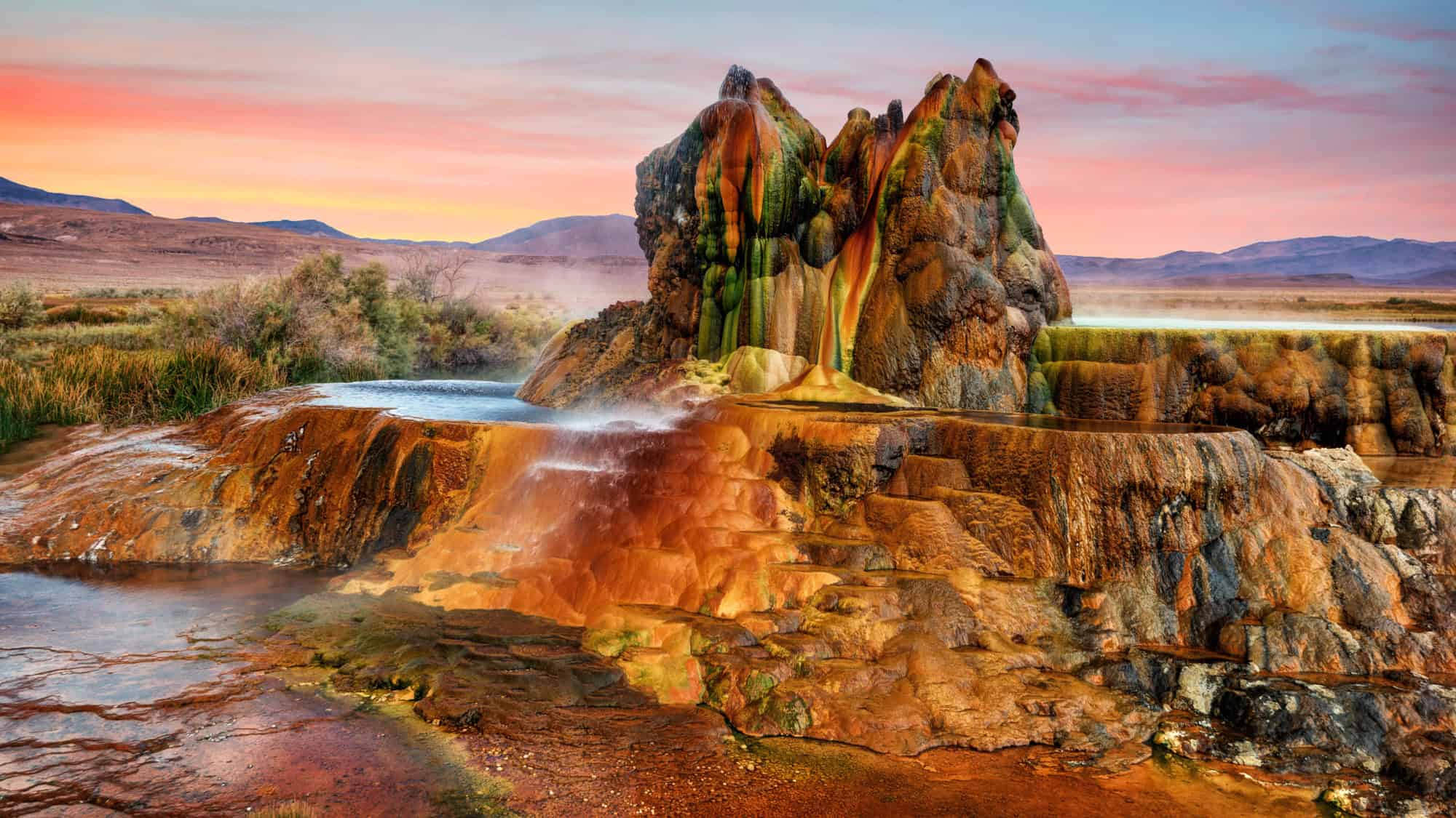 Colorful mineral terraces and vibrant geothermal formations of Fly Geyser erupt with steam under a pastel sunset sky in the Nevada desert.