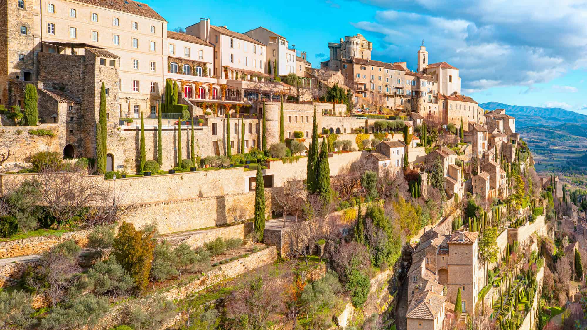 Hilltop village in southern France with terraced stone houses and narrow alleyways cascading down the hillside, bathed in golden sunlight and surrounded by lush greenery and distant mountain views.