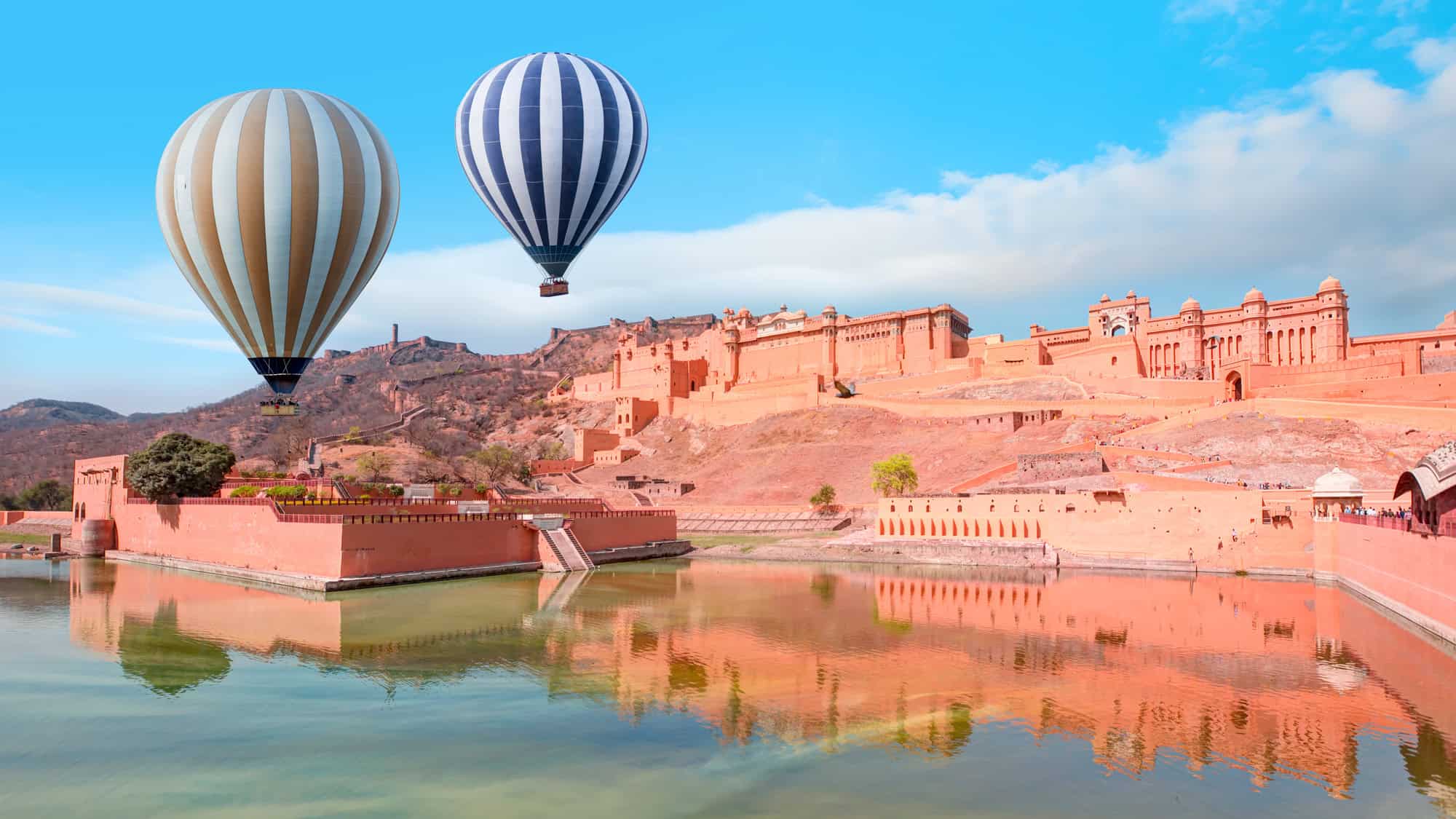 Two striped hot air balloons hover near a massive pink sandstone fort in Jaipur, India, with their reflections visible in the still water below the historic walls.