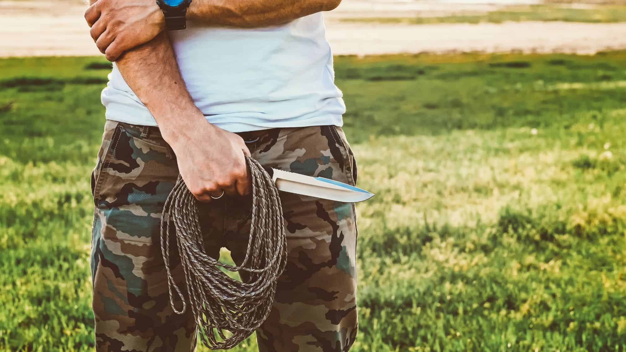 Close-up of a man wearing camo pants holding a coiled rope and a fixed-blade survival knife in a grassy open field, ready for outdoor tasks.