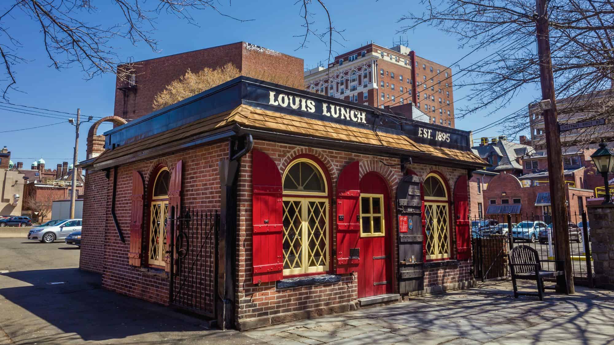 A historic brick building with red shutters and yellow-trimmed windows labeled “Louis Lunch Est. 1895” is seen under a blue sky, located on a street corner surrounded by other buildings.