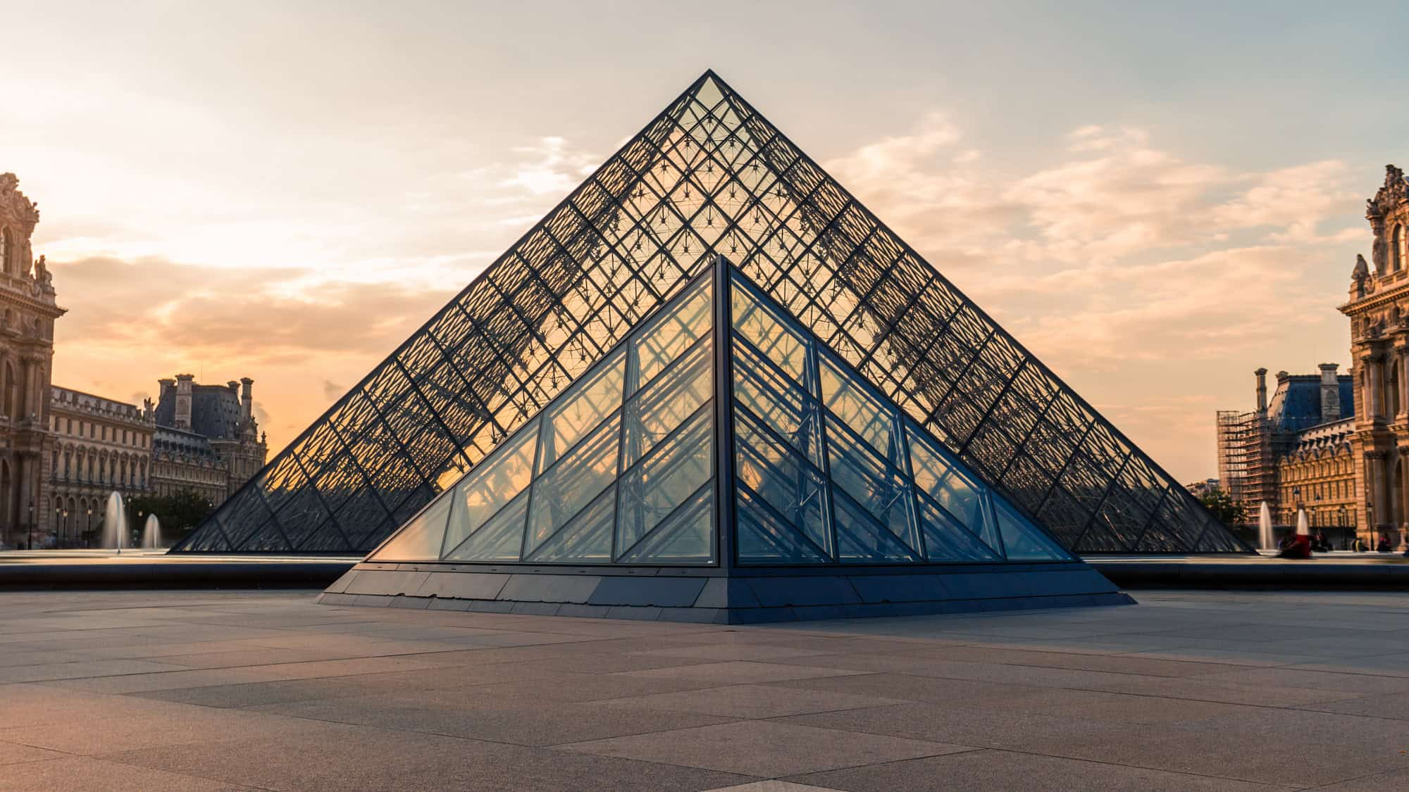 Modern glass pyramid of the Louvre Museum reflecting soft sunset light, flanked by classic French Renaissance architecture in the historic Parisian courtyard.