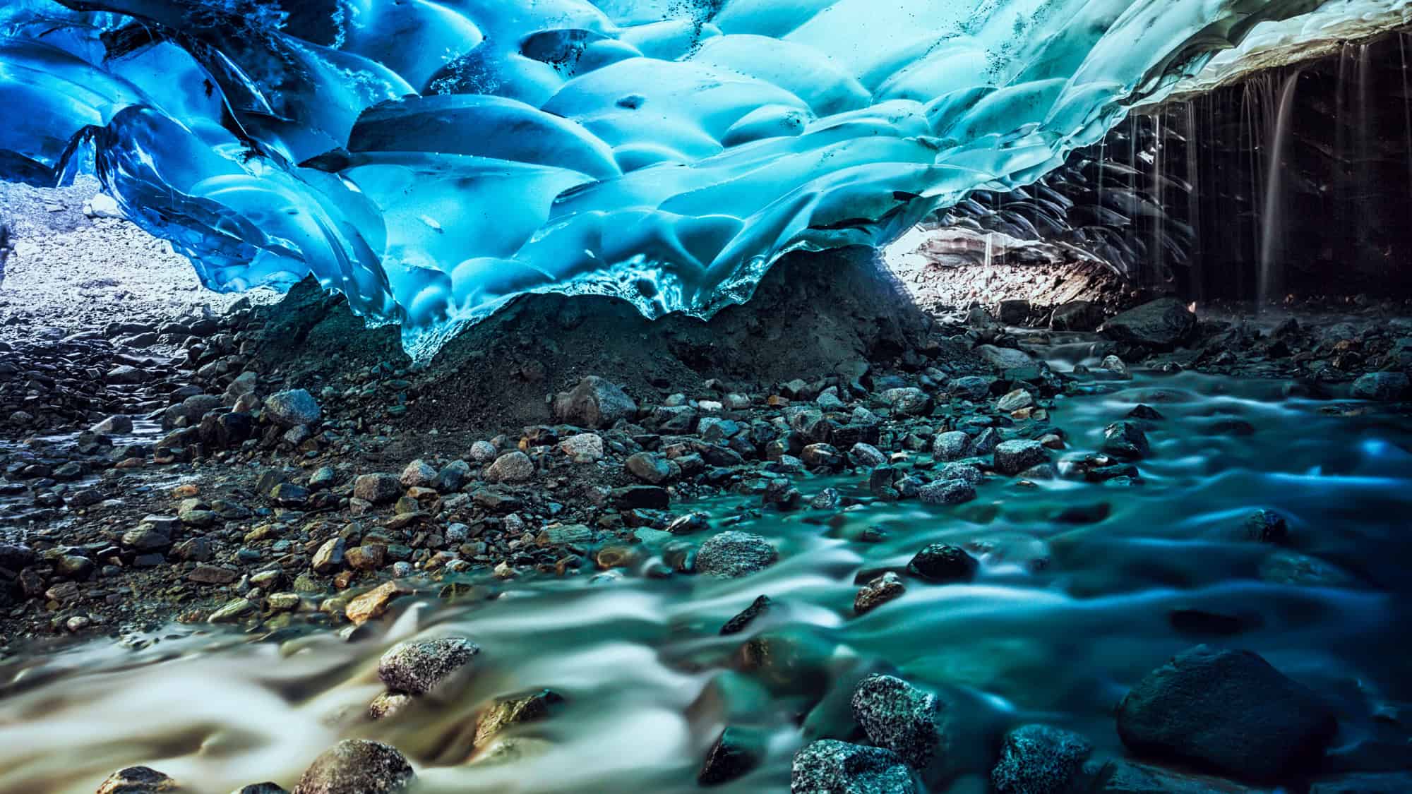 A surreal scene inside Alaska’s Mendenhall Ice Cave, showing glowing blue ice formations above a rocky floor and a gently flowing glacial stream.