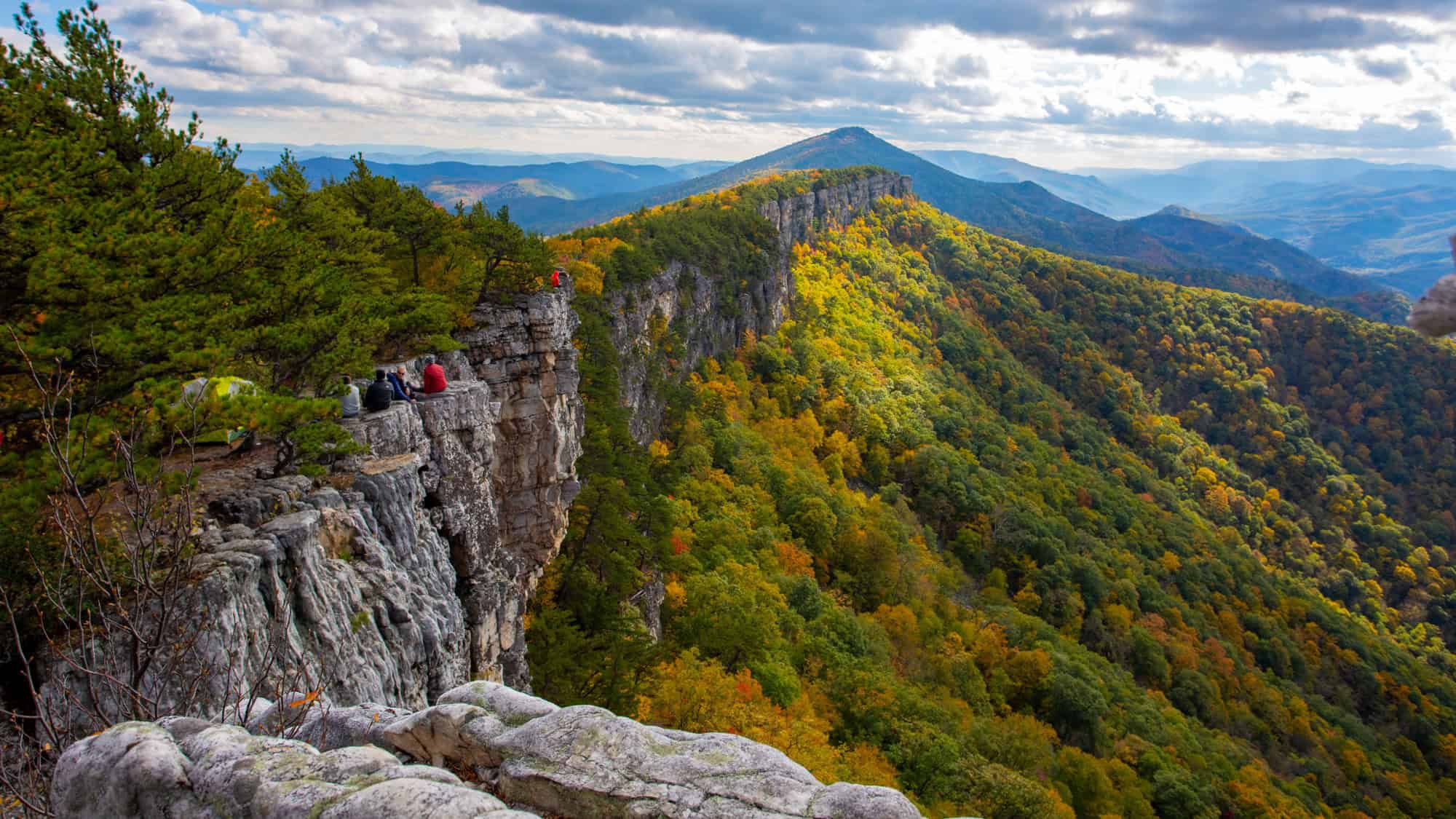 Hikers sit near the edge of a dramatic cliff, overlooking a stunning expanse of rolling mountains covered in colorful fall foliage beneath a partly cloudy sky.