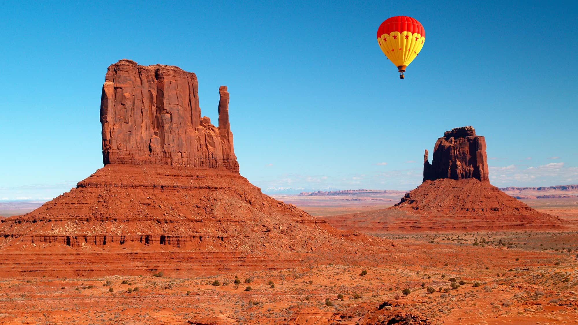 A vibrant red and yellow hot air balloon drifts between two towering red rock buttes in Monument Valley, Utah, beneath a deep blue sky.