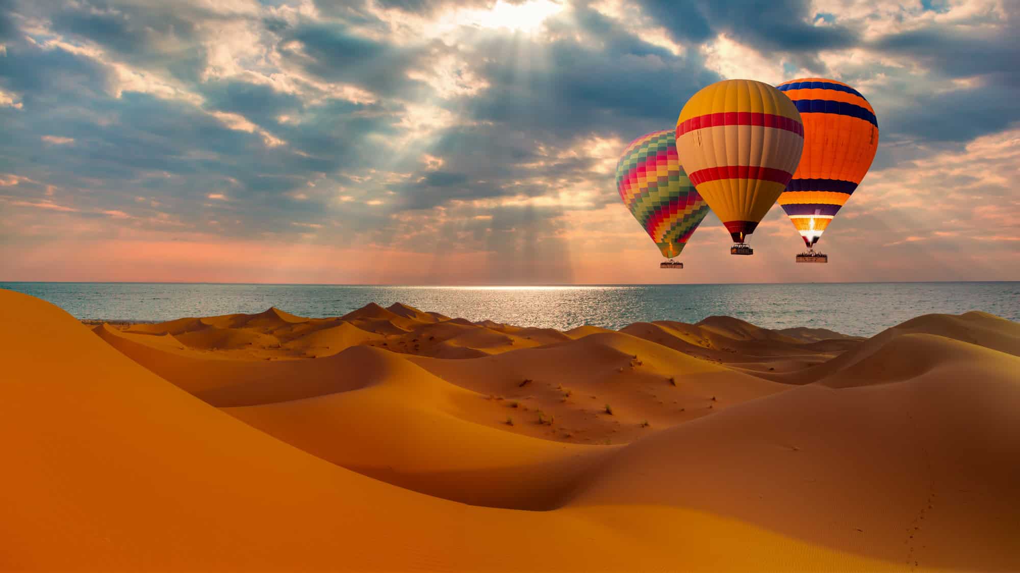 Three multicolored hot air balloons hover over rolling golden sand dunes beside the ocean in the Namib Desert, with sun rays breaking through dramatic clouds.