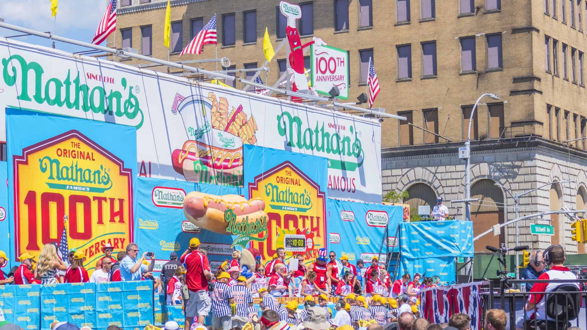 A vibrant street scene from Nathan’s Famous hot dog eating contest features large banners for the 100th anniversary and a crowd of spectators in red, white, and blue outfits.