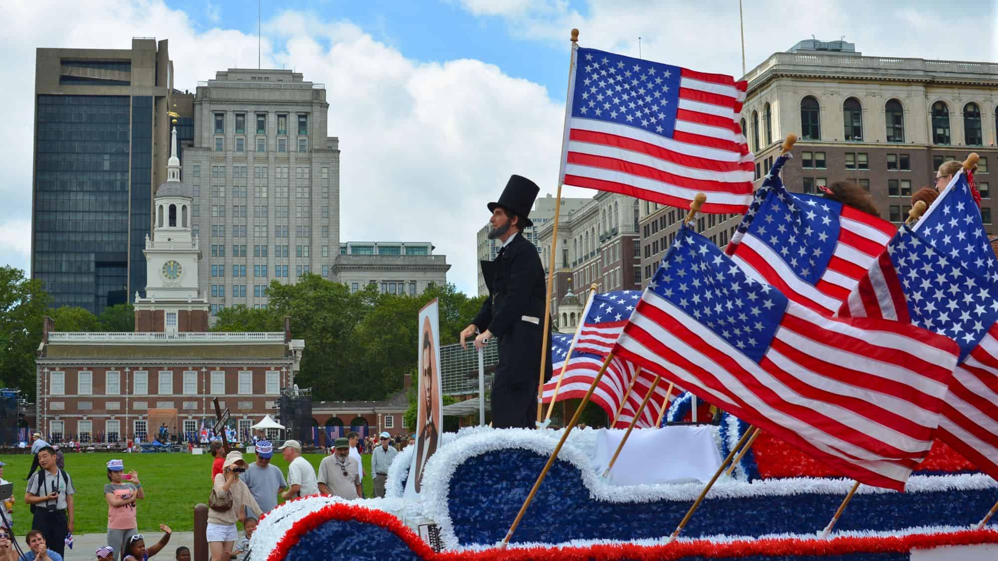 A float decorated in red, white, and blue tinsel carries an Abraham Lincoln impersonator and numerous American flags in front of Independence Hall, with a crowd gathered for a July 4th parade.