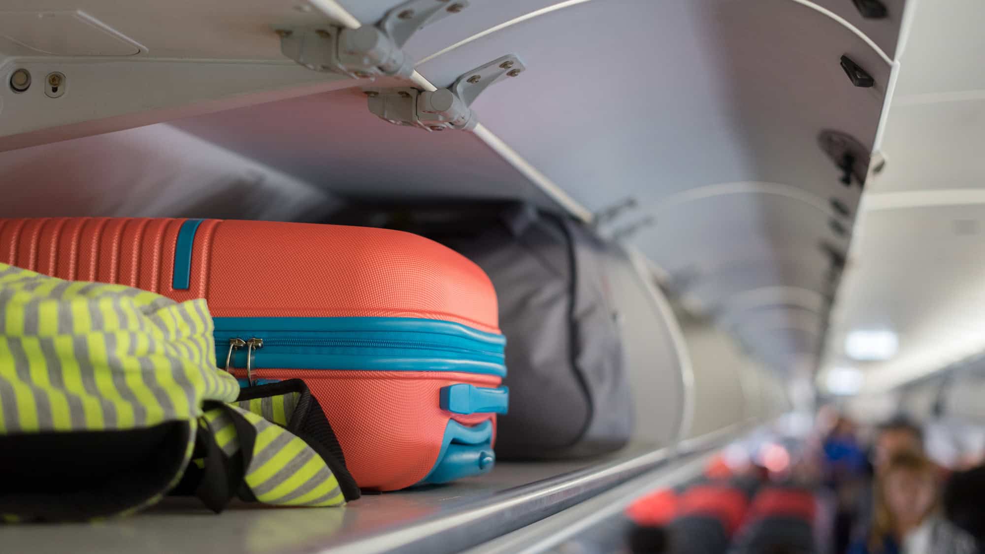 A close-up view of an airplane overhead bin filled with carry-on luggage, including a bright orange and turquoise hard-shell suitcase and a striped yellow backpack.