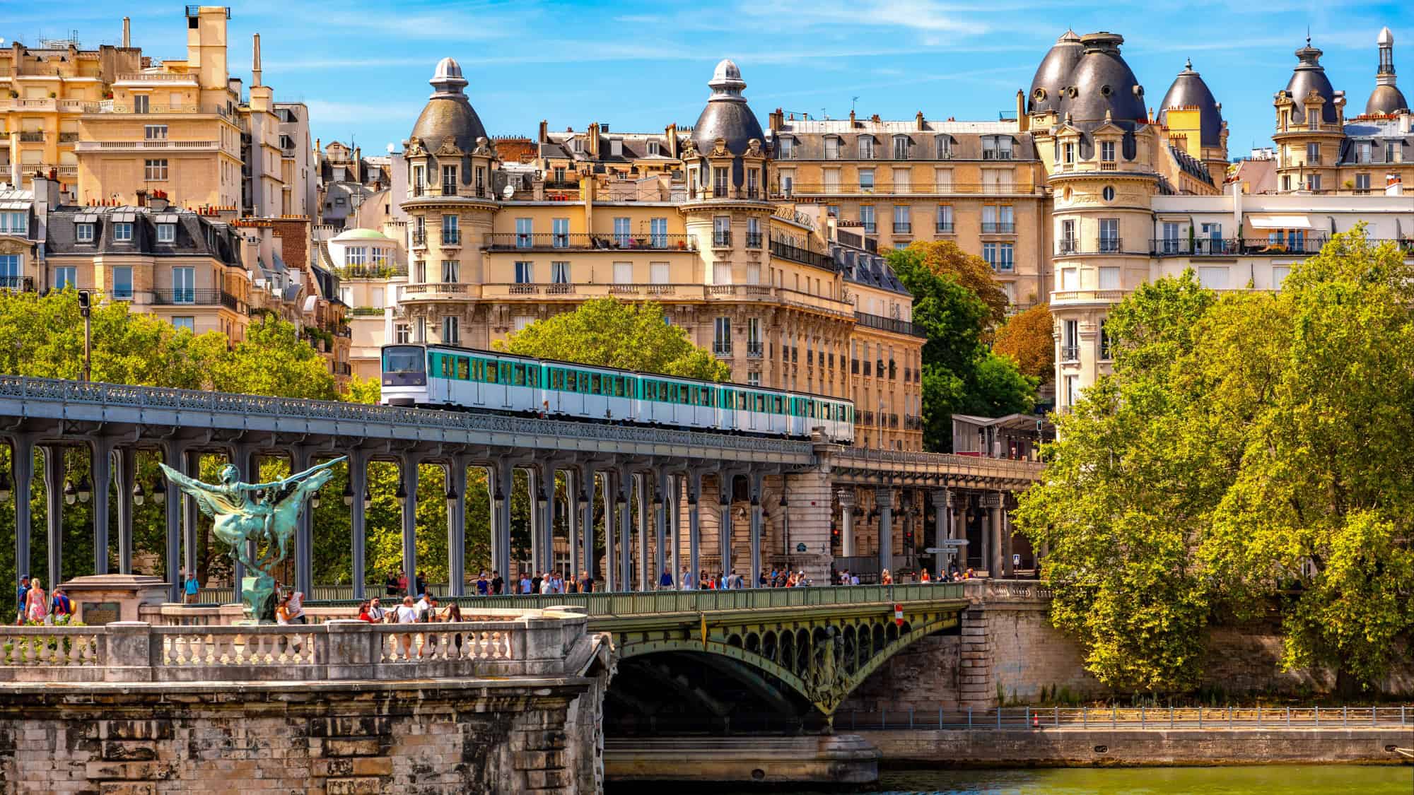 A green metro train crosses the Pont de Bir-Hakeim bridge in Paris above the Seine, surrounded by ornate buildings and leafy trees, with pedestrians strolling along the lower level.