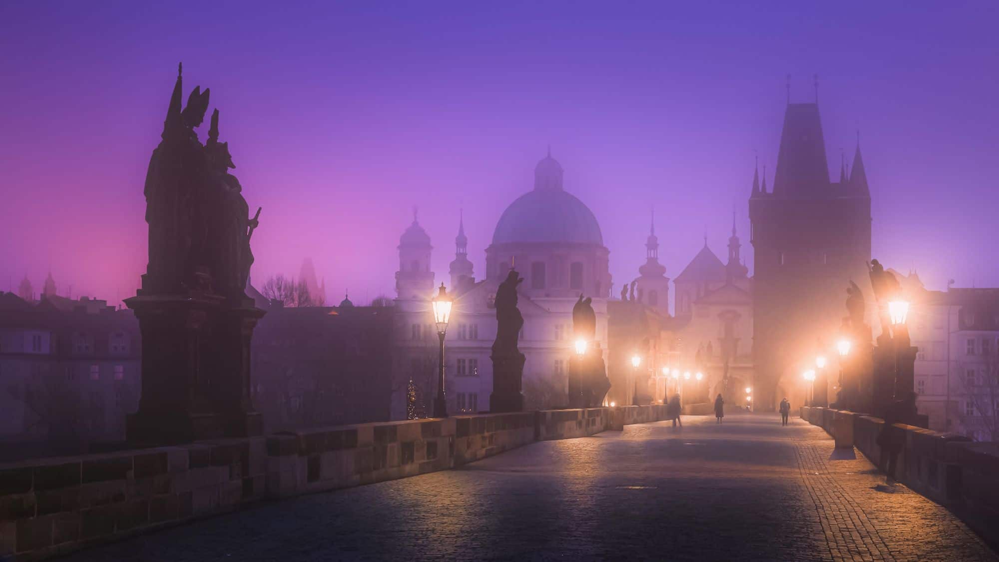 A dreamy purple fog blankets Charles Bridge in Prague at dusk, where silhouettes of historic statues and softly glowing lanterns line the cobbled path leading toward baroque architecture.
