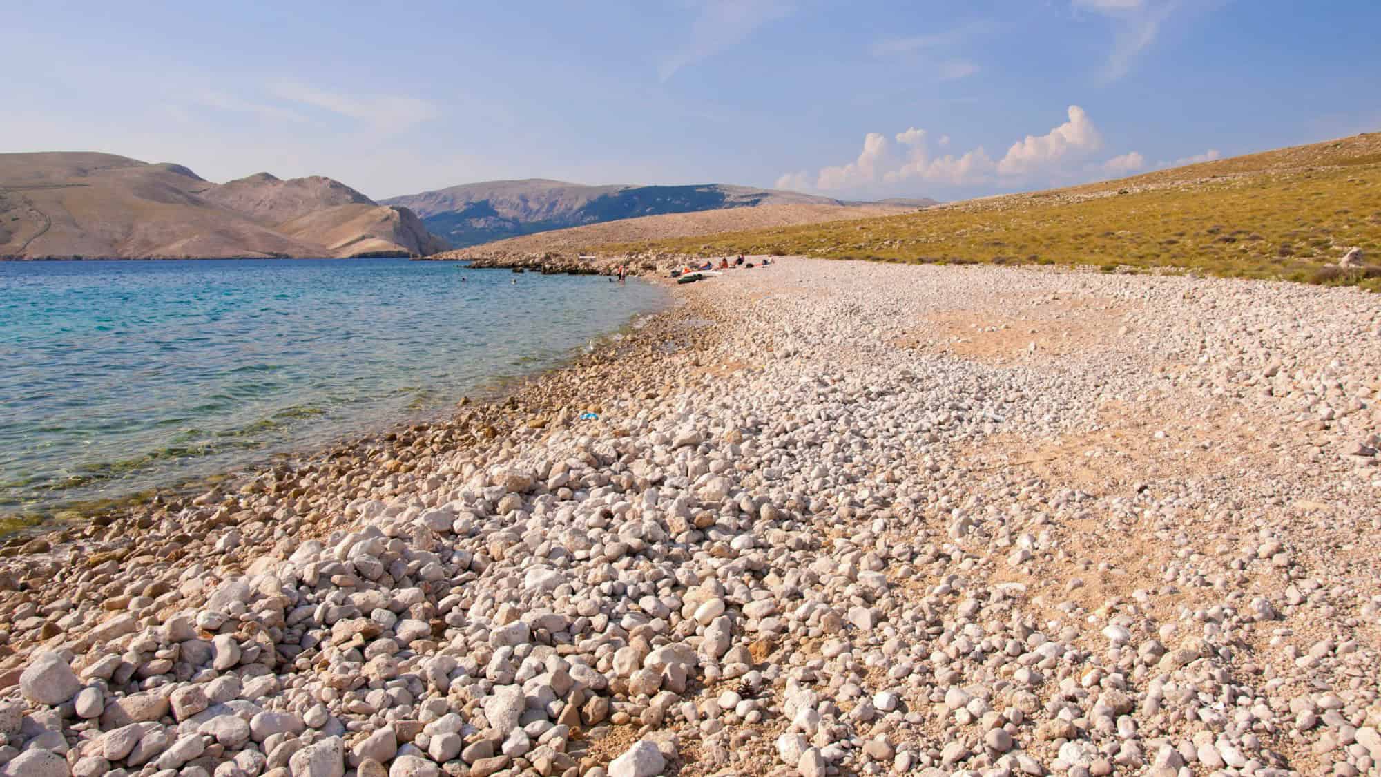 A rocky beach lined with smooth white stones slopes gently into the clear turquoise waters of the Adriatic, with arid, tan hills rising in the distance under a bright blue sky.