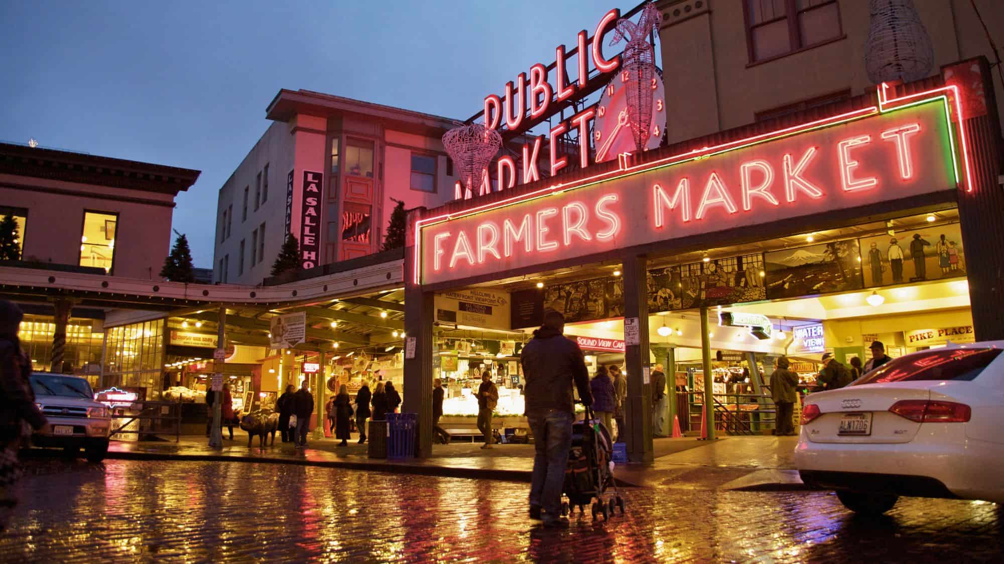 The neon “Public Market” and “Farmers Market” signs at Seattle’s Pike Place glow warmly at twilight, reflecting off the slick, rain-soaked brick street below.