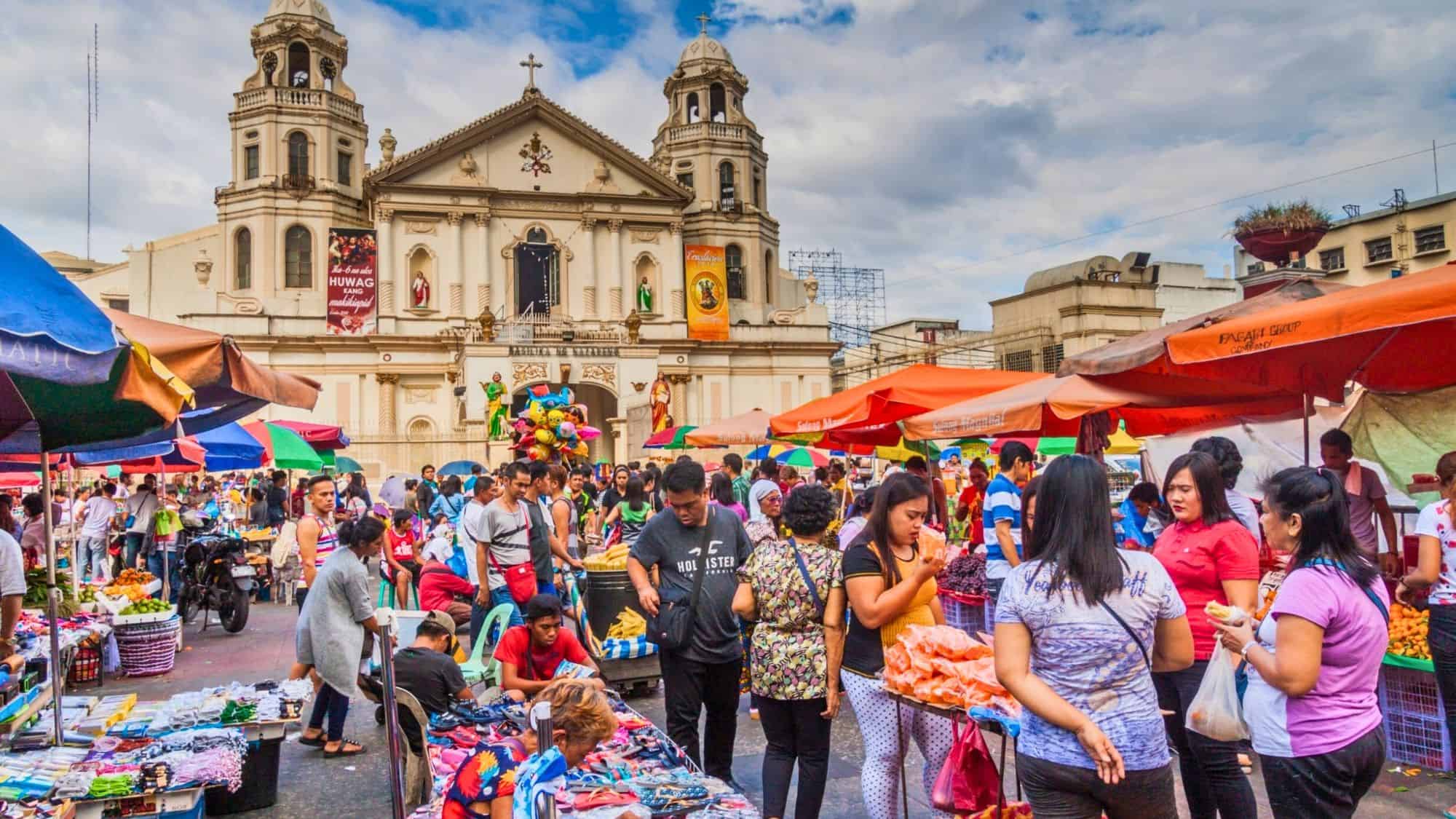 A bustling outdoor market scene in front of the Minor Basilica of the Black Nazarene in Quiapo, Manila, with vibrant vendor tents, crowds of shoppers, and colorful goods like clothes, fruits, and toys on display.