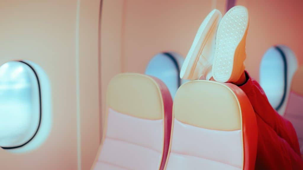 A person wearing white sneakers and red pants has their feet propped up against the back of an airplane seat, showing poor in-flight etiquette.