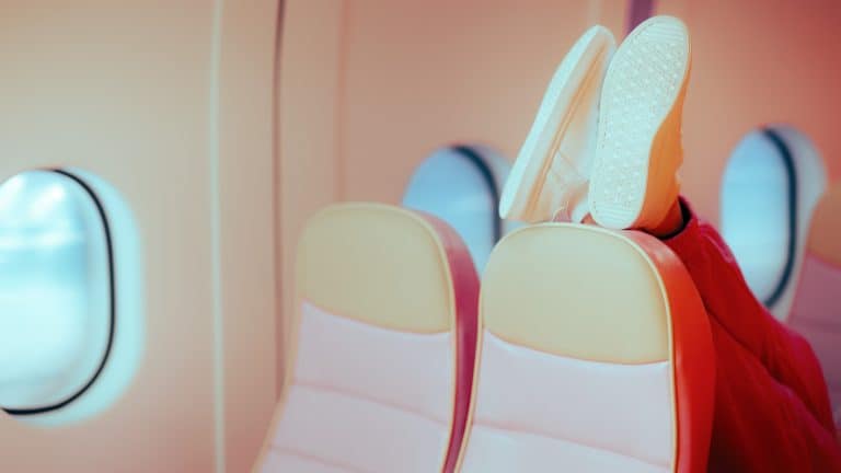 A person wearing white sneakers and red pants has their feet propped up against the back of an airplane seat, showing poor in-flight etiquette.