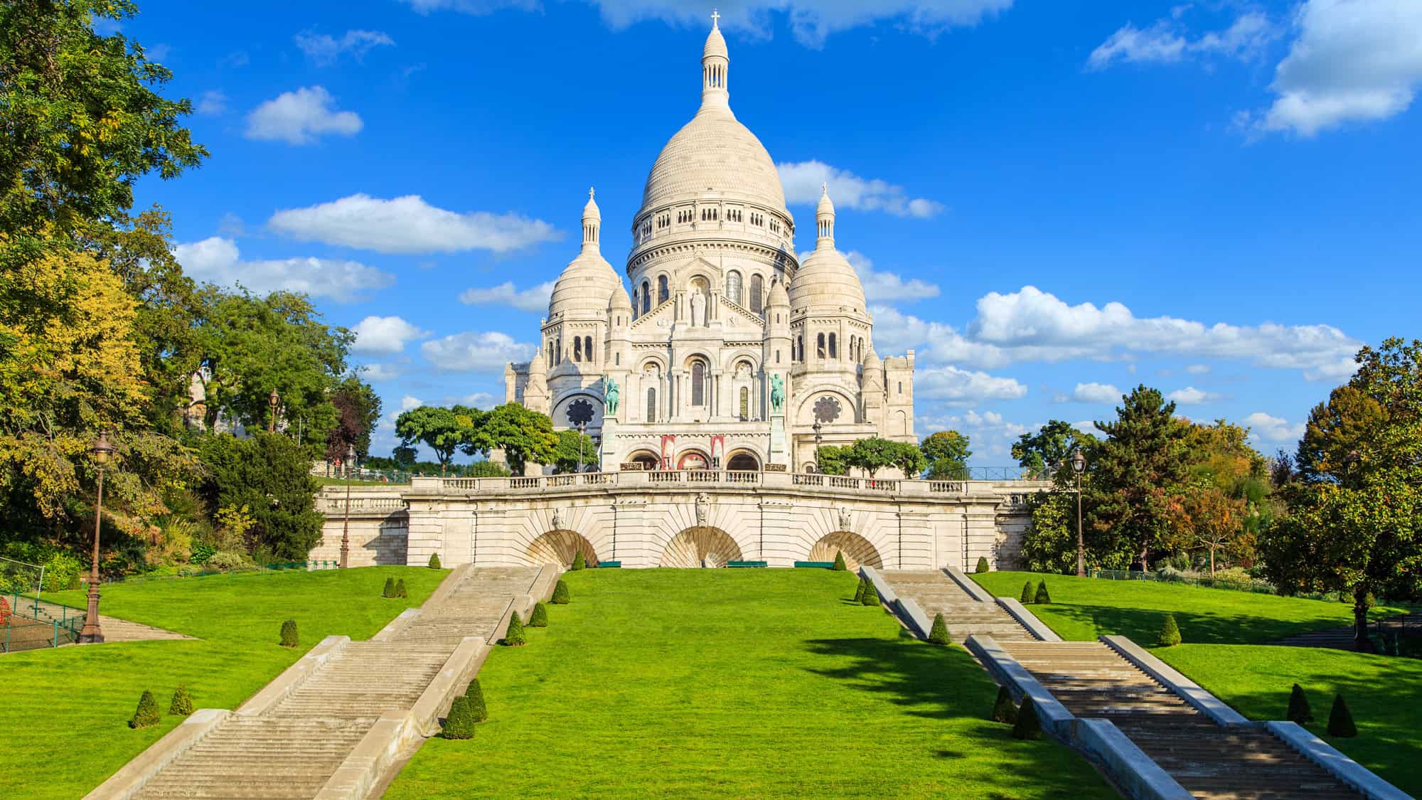 Bright white domed basilica of Sacré-Cœur sits atop Montmartre in Paris, framed by green lawns and symmetrical stairways under a clear blue sky.