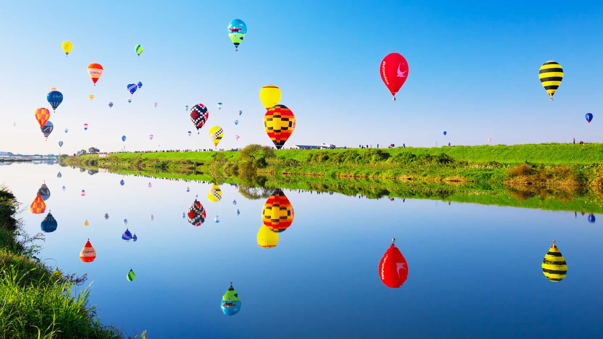 Dozens of hot air balloons in a variety of colors and patterns are launched along a calm river, with their reflections perfectly mirrored in the glassy surface, surrounded by green embankments and clear blue skies.